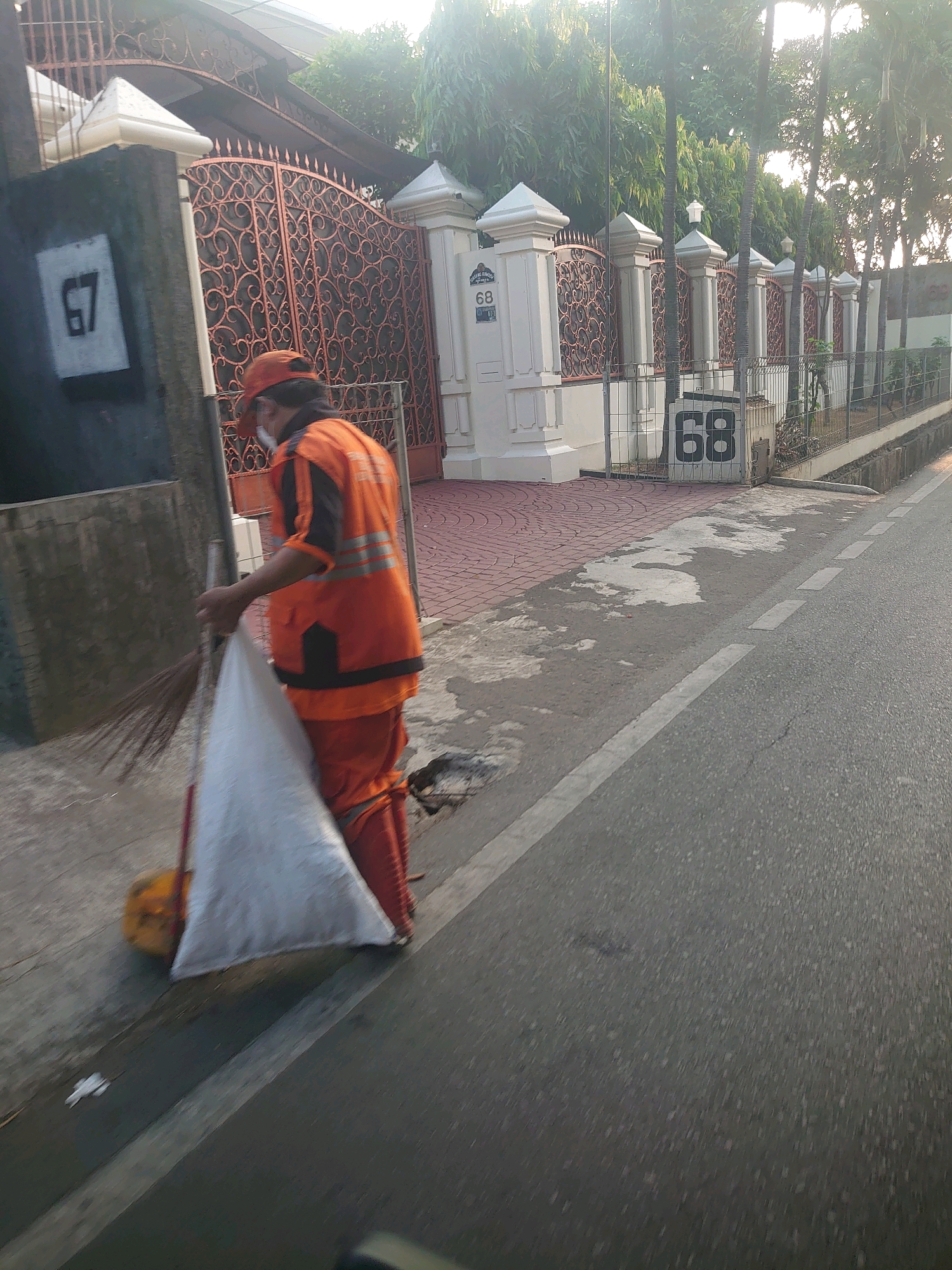 A street cleaner in an orange uniform and cap is sweeping debris on a sidewalk next to a fence and a building. The cleaner holds a broom in their right hand and is pushing a large white bag filled with debris with their left. The bag rests on a round yellow object. The sidewalk is paved with red brick and gray concrete with patches of water. The fence is made of metal with intricate scrollwork and is supported by white pillars. House numbers "67" and "68" are visible on the wall and gate. The street is asphalt with white lane markings. The background shows lush green trees and palm trees. The scene is illuminated by the warm light of sunrise or sunset, casting long shadows. The overall impression is of a quiet morning in a residential area of South Jakarta, Indonesia.