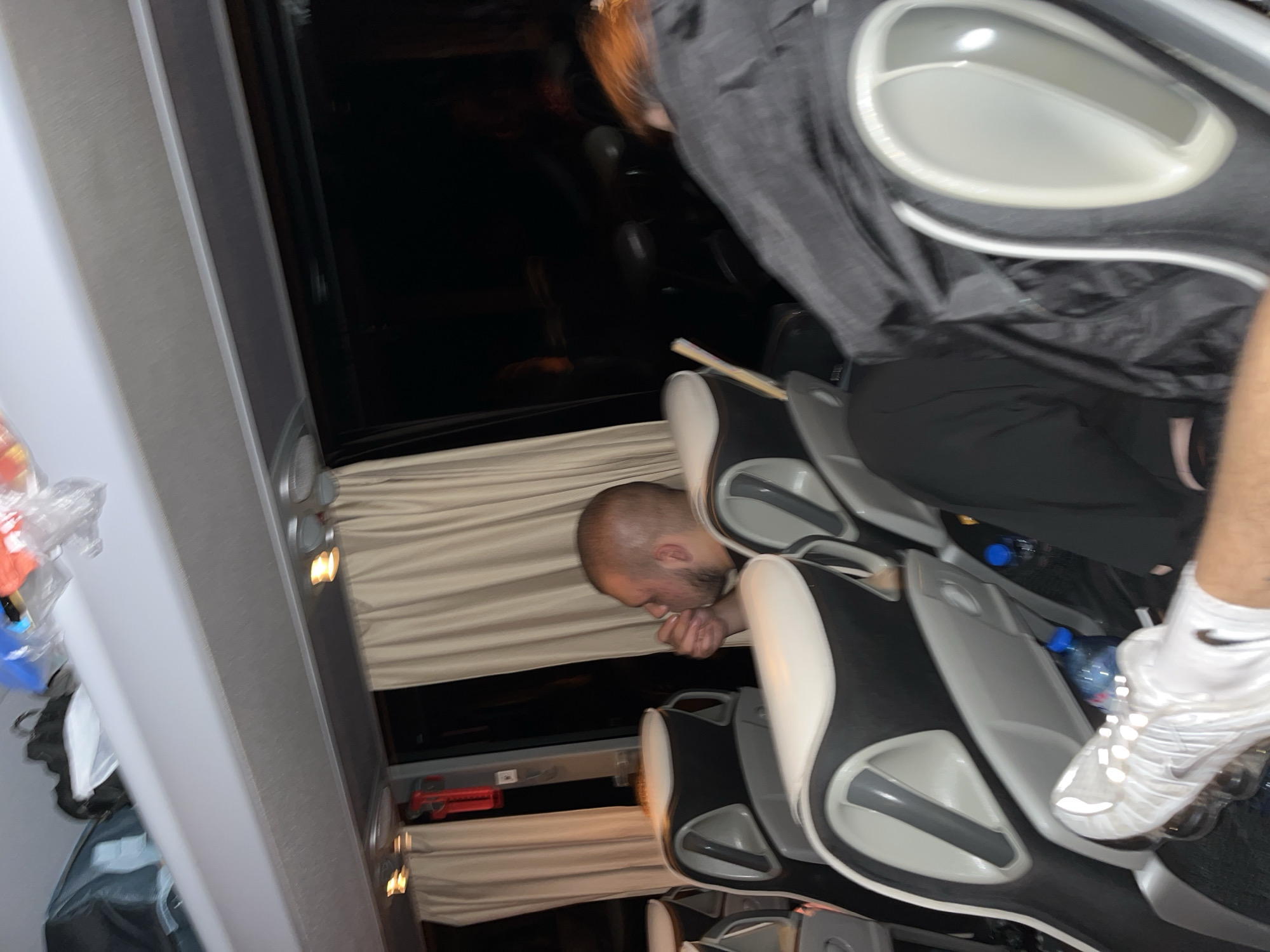 A man with a shaved head is visible through a bus window. He has his hand on his chin, and appears to be sleeping. The bus seats are grey with white headrests. A dark curtain covers the window behind him. There are two plastic water bottles on the seats. The bus is traveling in Milevo, Bulgaria.