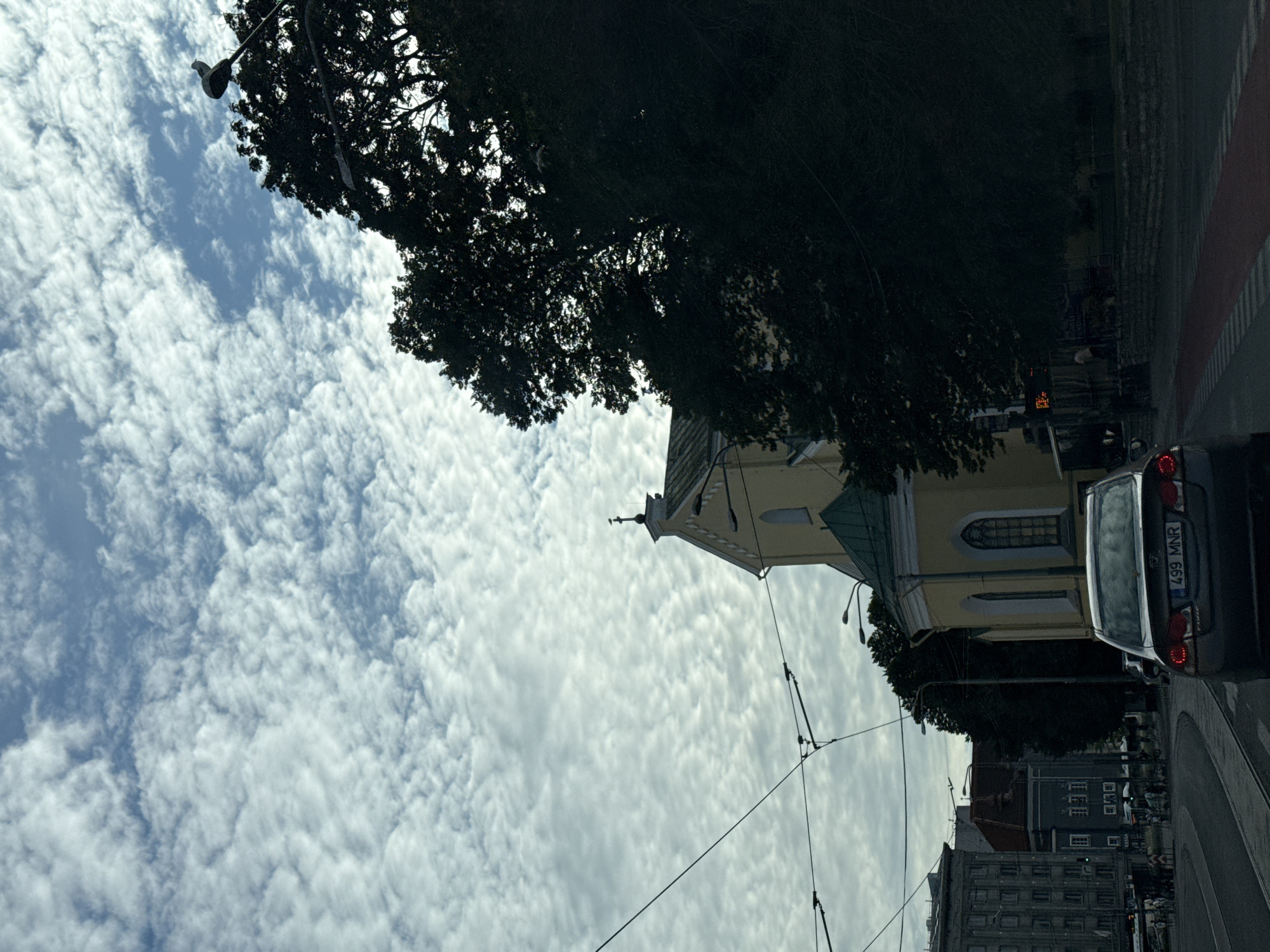 The image depicts a street scene in a city, under a sky filled with scattered, wispy clouds. In the foreground, a dark-colored car with a license plate reading "499 MNR" is visible from the rear, positioned on a road with tram tracks. To the right of the car, a yellow building with a green roof, possibly a church, is partially obscured by the dense foliage of a large, dark tree. The building features arched windows and a cross at its apex. Further to the left, other city buildings are visible, along with overhead power lines for what appears to be a tram system. Pedestrians are present on the sidewalk near some of these buildings. Streetlights and trees line the road, contributing to the urban environment.