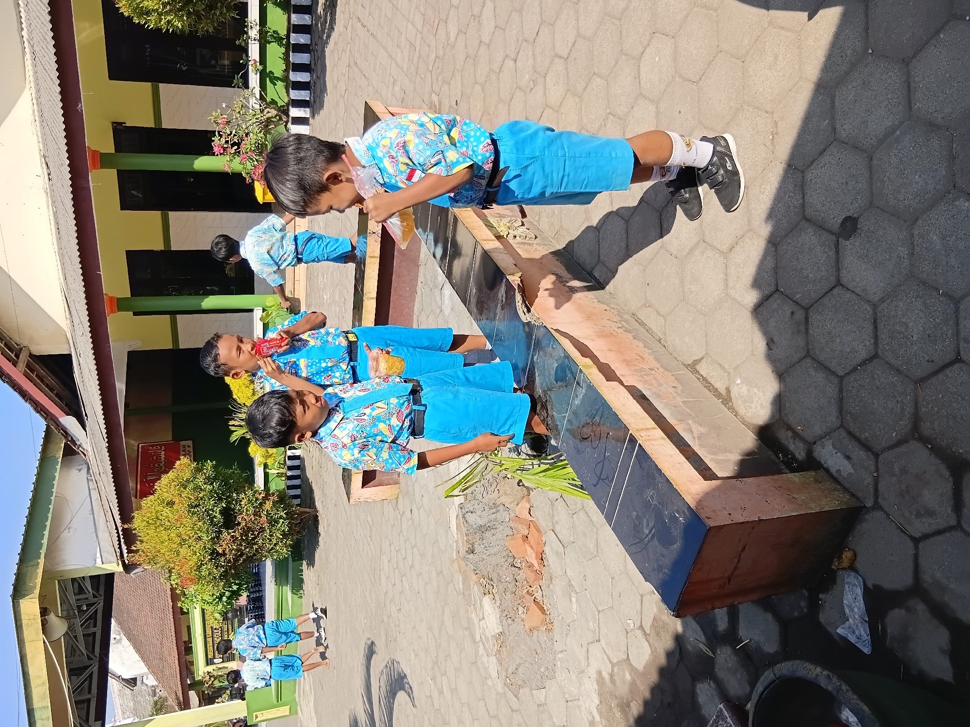 In Tanggul, Indonesia, three young boys in blue school uniforms are standing on a paved courtyard. The boy on the left is looking forward, while the boy in the center is holding a red candy bar to his mouth. The boy on the right is drinking from a clear plastic bag. Behind them, a low wall runs across the courtyard, and a building with a yellow facade and green trim is visible. The ground is covered in hexagonal paving stones, and there is a trash can on the left. The scene appears to be a schoolyard during the day.