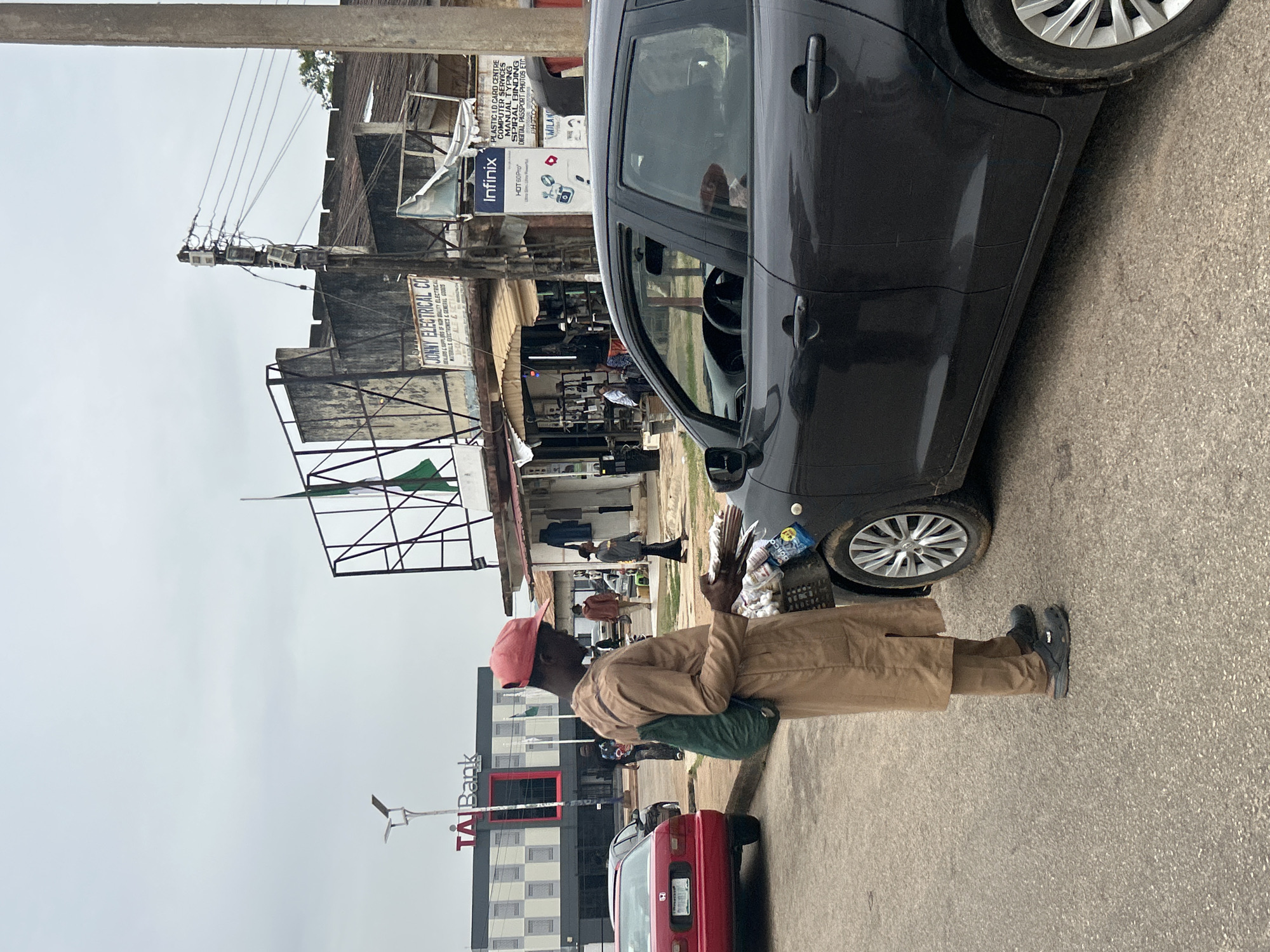A medium, angled shot depicts a street scene in Jos, Nigeria.
Foreground: A black sedan is partially visible in the upper right corner, positioned on the roadside. Standing beside the car is a dark-skinned man wearing a pink cap, tan jacket, and dark shoes. He carries what appears to be goods or wares. A red sedan is slightly behind him.
Background: Buildings and commercial establishments line the street. Notable signs include "TAI Bank," "JONNY ELECTRICAL CO," and "Infinix" advertising electronics. Electrical wires run overhead. Other people are visible walking along the street. The sky is overcast.