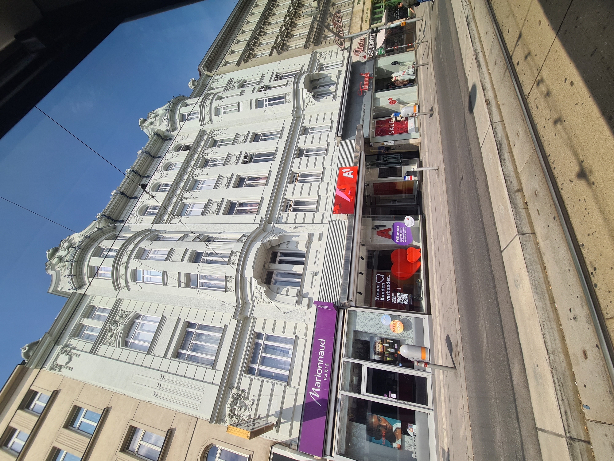 The image captures a street scene in Vienna, Austria, focusing on the facade of several buildings. On the left, a pale yellow building with classical architectural details is visible, followed by a light green, ornate building with a curved bay window. This building houses several shops, including "Marionnaud PARIS" with a prominent purple awning, and "A1," an Austrian telecommunications company, indicated by its red and white logo. Further to the right, a shop with a sign that reads "Triumph" and another with "Oida Cafe-Konditorei" are seen, followed by a building with the text "THE TASTE OF ASIA" and what appears to be a green awning. The street in front of the buildings is paved with asphalt, featuring tram tracks and a concrete sidewalk. Several street poles, some with signage, are present along the sidewalk. The sky is clear and blue. The perspective is from a moving vehicle, likely a tram, looking out at the street.