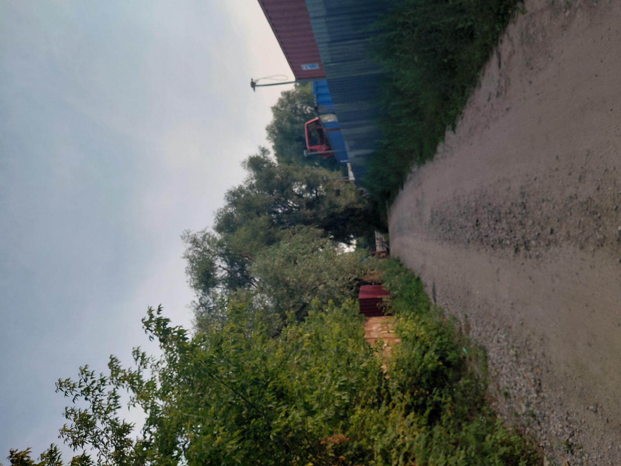 The image depicts a dirt road in an industrial or semi-urban area, likely on the outskirts of Novosibirsk, Russia. On the left side of the road, lush green trees and bushes line the path, with a red fence partially visible behind them. On the right side, a corrugated metal fence separates the road from a yard containing several large shipping containers, one of which is red and the other blue. A red vehicle chassis, possibly from a truck or van, is visible atop the blue containers. A tall, thin lamppost with a coiled wire extends upwards from behind the fence. The sky is overcast with a pale, grayish-blue hue. The road itself is unpaved, covered in gravel and dirt, suggesting it is not a main thoroughfare. The overall impression is one of a functional, somewhat neglected, working area.