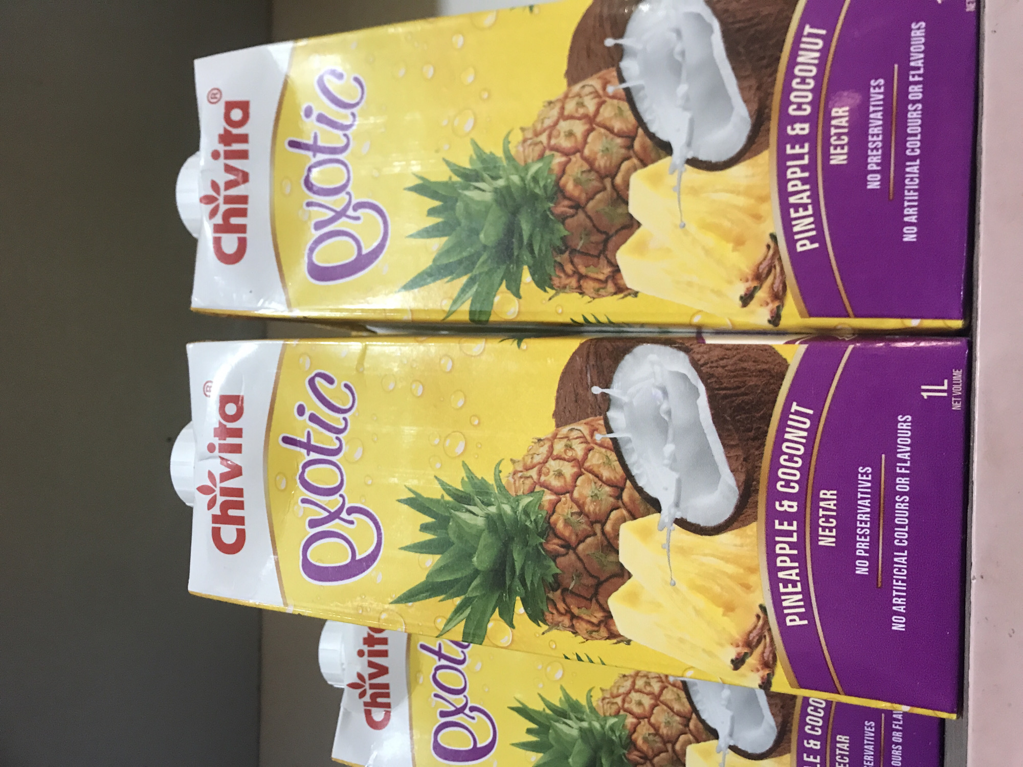 Two Chivita Exotic Pineapple & Coconut Nectar cartons are on display, side-by-side on a shelf, likely in a retail setting in New Stadium, Nigeria. The cartons have a vibrant yellow background with water droplet graphics. Prominently featured are images of pineapples and coconuts, along with the text "Chivita," "Exotic," "Pineapple & Coconut Nectar," "No Preservatives," and "No Artificial Colours or Flavours." One carton is more visible than the other, with the left carton partially obscured. The bottom of the cartons indicate a net volume of 1L. The lighting suggests an indoor environment.