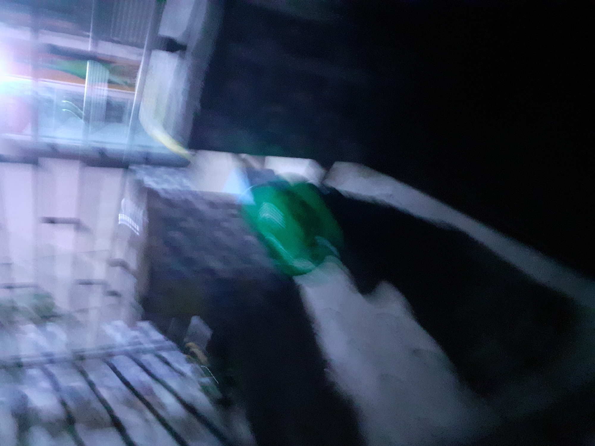 The image depicts a blurry, low-light scene of an outdoor area, likely a courtyard or patio, in Jalingo, Nigeria. In the center of the frame, a bright green object, possibly a watering can or a bucket, is visible and appears to be in motion, suggested by motion blur. The background features architectural elements, including what seems to be a brick wall or structure on the left, with horizontal lines suggesting shelving or stacked items. To the right, there is a dark, rectangular object, likely a container or furniture, and further back, a blurred background with vertical lines and a bright light source, possibly a window or an illuminated opening. The ground appears to be paved or made of concrete. The overall impression is of a candid, possibly nocturnal shot, characterized by significant motion blur and limited detail due to poor lighting conditions.