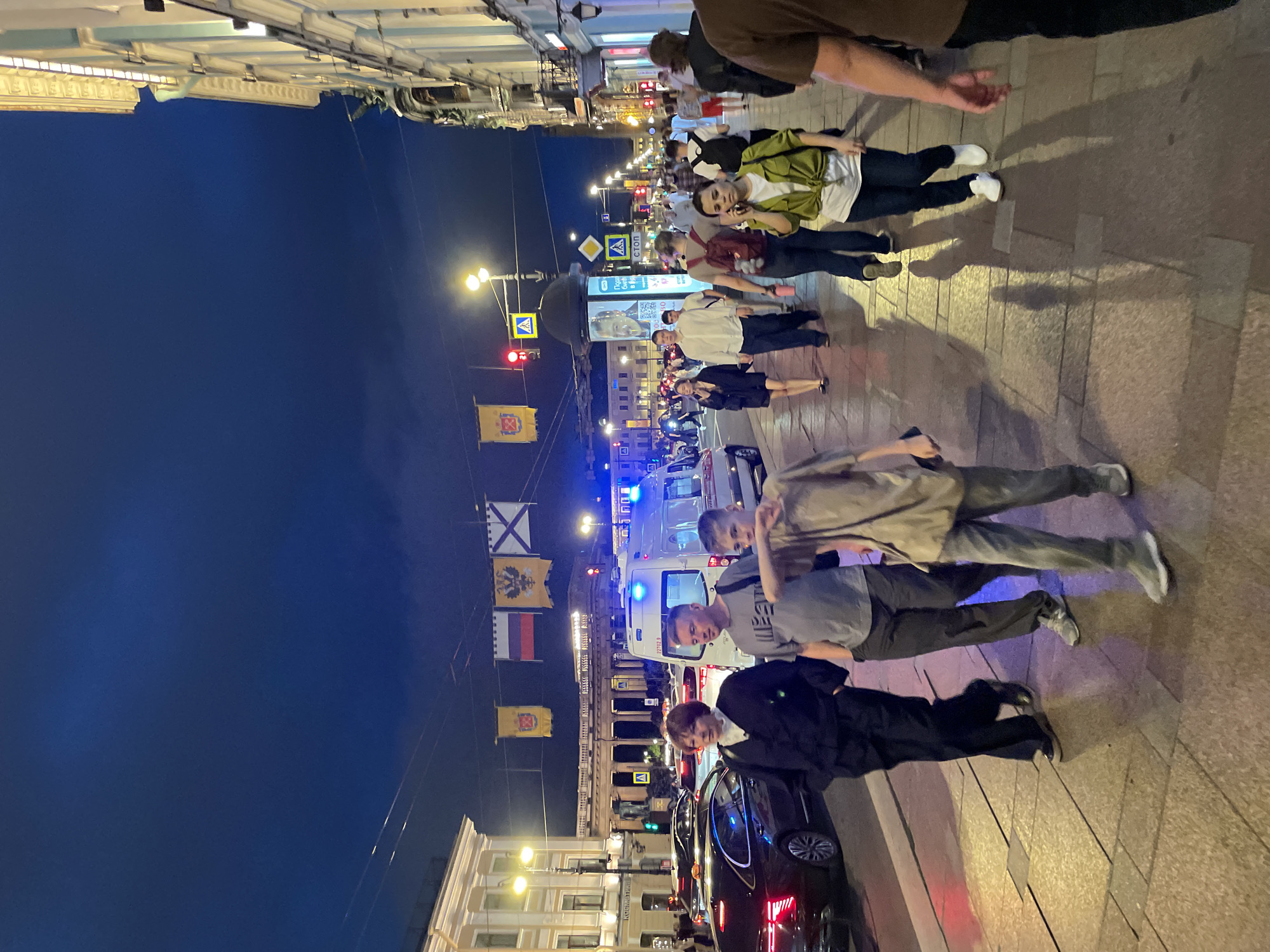 This image captures a bustling evening scene on Nevsky Prospekt in Saint Petersburg, Russia, looking towards the Kazan Cathedral, whose grand colonnade is visible in the background under a dark blue night sky. Overhead, four distinctive flags are strung across the street: a yellow flag bearing the coat of arms of Saint Petersburg (crossed anchors and a scepter), the Russian national flag (white, blue, red horizontal stripes), a yellow flag with a black double-headed eagle (imperial standard), and a white flag with a blue diagonal cross (St. Andrew's flag).
The wide street accommodates both vehicular traffic and numerous pedestrians. On the left, several cars are visible, including a dark sedan with illuminated red taillights. A white ambulance with activated blue flashing lights on its roof and sides is prominent in the central roadway. The scene is illuminated by multiple artificial light sources, including streetlights, vehicle headlights and taillights, and lights from buildings and an advertising kiosk.
The foreground features a wide, light-colored paved sidewalk occupied by many pedestrians. Three individuals are particularly distinct: a woman with short reddish-brown hair wearing a dark blazer over a white high-collared shirt and dark trousers; a man with short brown hair wearing a grey t-shirt, dark trousers, and carrying a dark shoulder bag; and a boy with light brown hair wearing a light brown t-shirt and light grey trousers, walking with his right arm bent and slightly raised. Other pedestrians are visible further down the sidewalk, engaged in walking and casual interaction.
The environment includes classical-style buildings lining both sides of the street, some with illuminated ground-floor establishments. Street furniture includes tall lampposts, traffic lights (one red, one green), pedestrian crossing signs, a "СТОП" (STOP) sign, and a cylindrical advertising column with an illuminated display showing a man's face. Shadows of pedestrians are cast on the illuminated pavement, creating a dynamic effect.