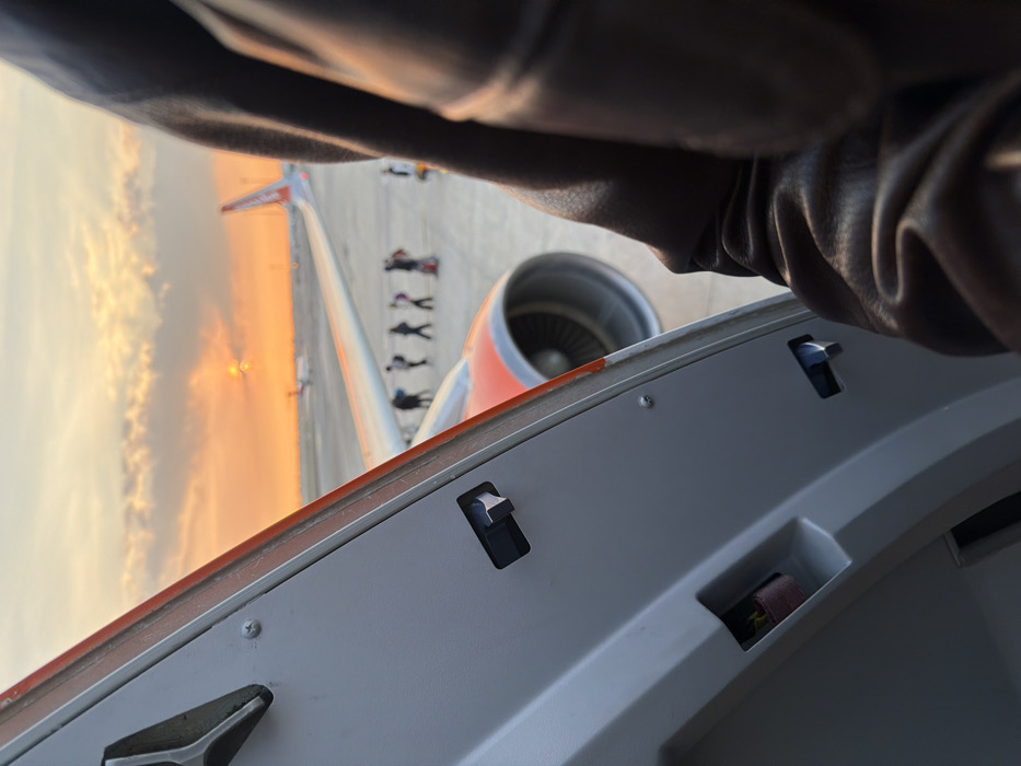 The image depicts an airport tarmac scene at sunset, observed from within an aircraft at Bordeaux-Mérignac Airport (BOD) in Mérignac, France. The perspective is through an open aircraft door or large window, with the light grey interior paneling, including a black latch mechanism and fasteners, visible on the left. A dark brown, textured object, likely a jacket sleeve or gloved arm, partially obstructs the right side of the view.
Immediately outside the aircraft, the white wing with an orange stripe and the front of a jet engine nacelle, showing fan blades, are visible. On the grey asphalt tarmac below, a line of approximately six to seven individuals, dressed in dark clothing, are walking in single file from left to right across the frame. Further to the right, another individual or small group is present. In the mid-distance, the tail section of an aircraft with an orange vertical stabilizer, identifiable as an EasyJet livery, is visible. Other indistinct aircraft and airport infrastructure are present on the distant tarmac.
The sky is illuminated by a sunset, exhibiting strong orange and yellow hues near the horizon, transitioning to lighter tones above. Scattered dark clouds are present. The setting sun is a bright, obscured orb near the horizon line. The overall lighting is warm, indicative of golden hour conditions.