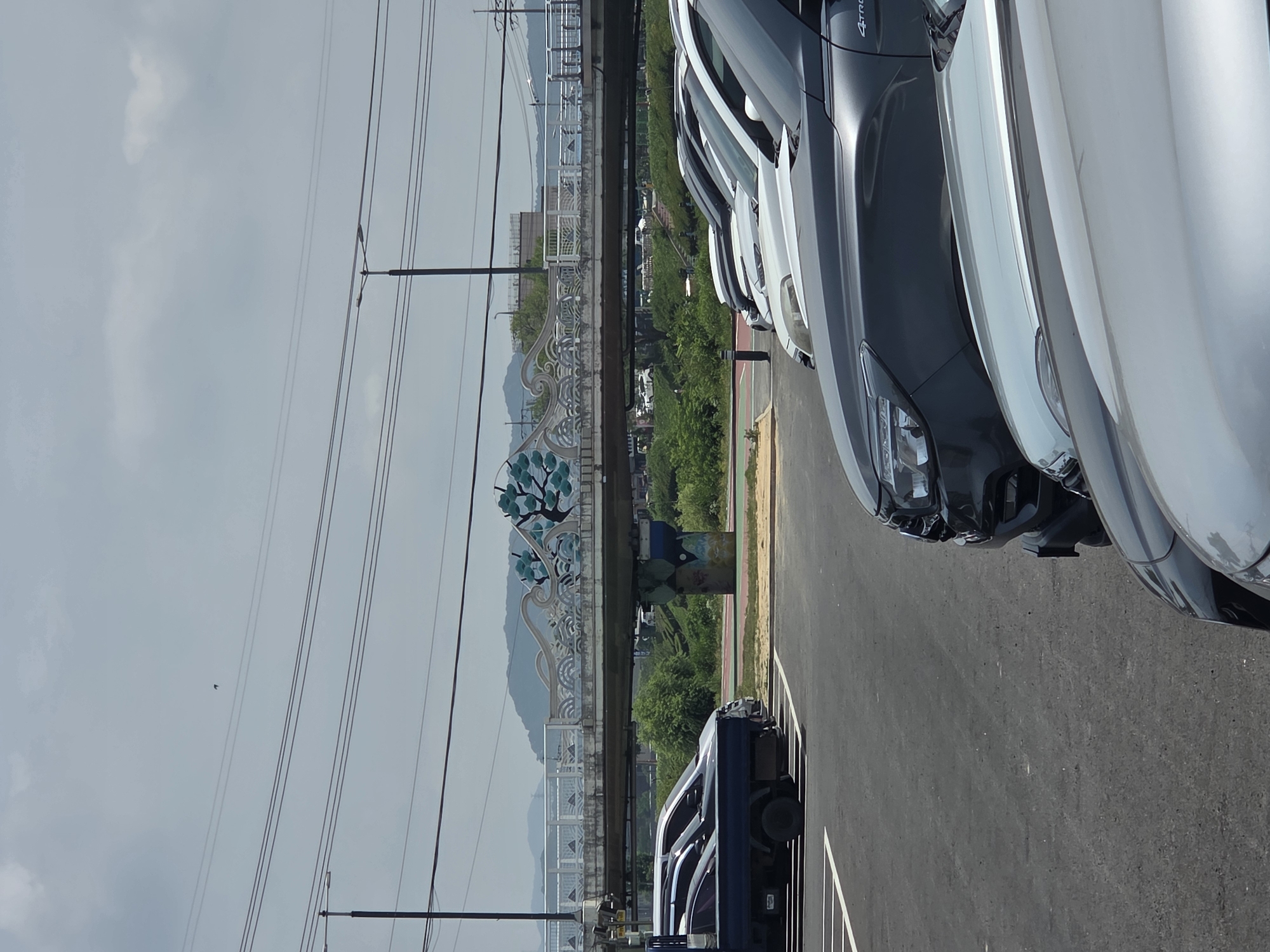 A parking lot is shown in front of a pedestrian bridge in Anseong-si, South Korea. Several cars, including a dark blue truck, a white SUV, and a dark grey SUV, are parked. The bridge has ornate white railings and a decorative element featuring a stylized tree with teal leaves. The bridge spans over a green, vegetated area with a road or path visible below. In the background, mountains are visible under a cloudy sky, with power lines running overhead. A single bird is visible in the sky. The scene appears to be captured on a daytime.