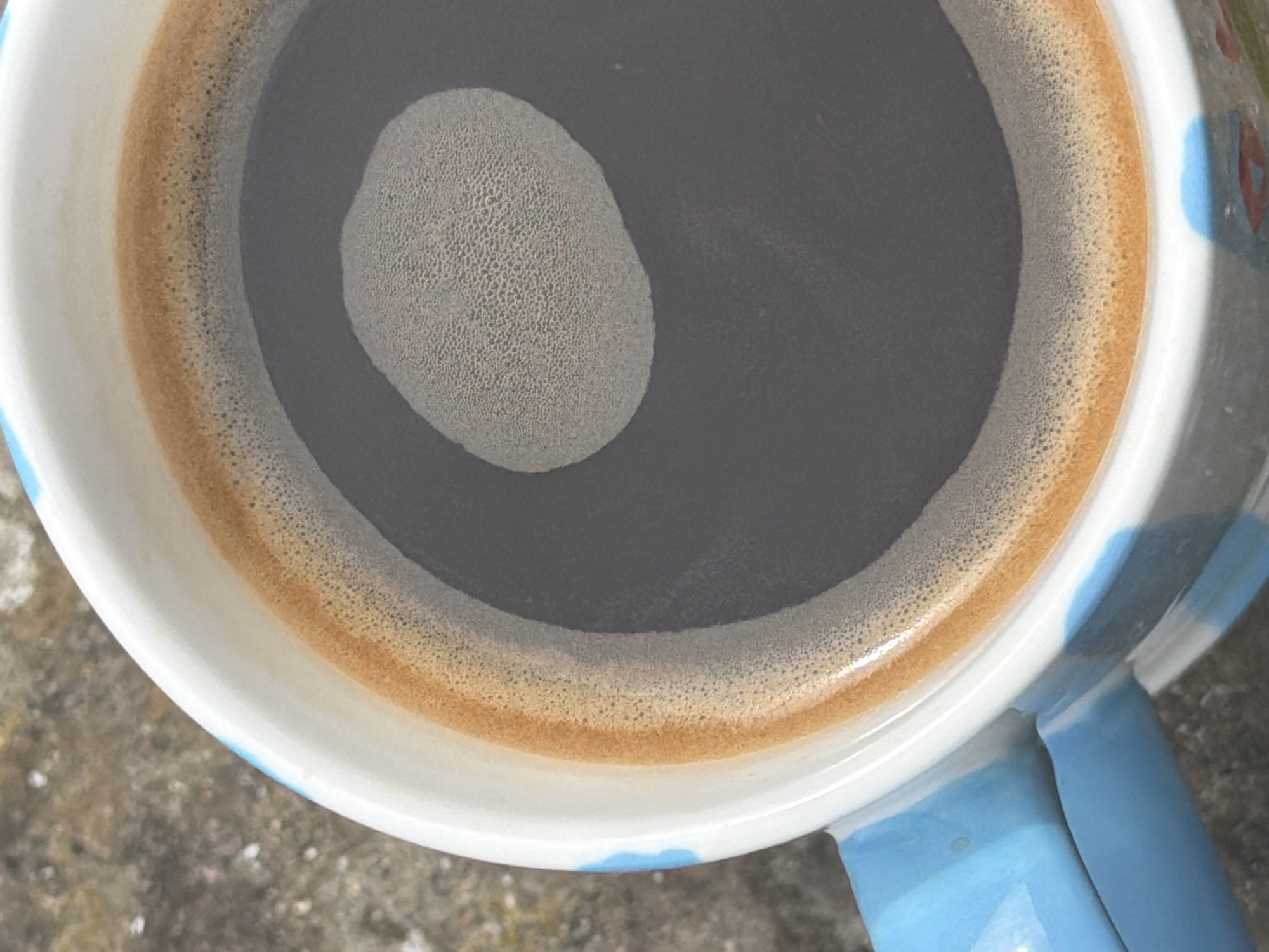 The image presents a close-up, top-down view of a mug of coffee. The coffee itself is dark and black, filling the mug. A distinct, reddish-brown ring of crema, characterized by a fine, frothy texture of numerous small bubbles, encircles the interior edge of the mug. Within the dark liquid, a prominent oval-shaped patch of lighter, denser foam, also exhibiting a very fine, intricate bubbly texture, floats centrally.
The coffee is held in a white ceramic mug that features blue accents, including a visible handle on the right side of the frame and what appears to be part of a blue and red patterned design on its exterior, seen near the rim. The mug is placed on a rough, grey surface, which resembles concrete or stone, suggesting an outdoor or unpolished setting. The lighting is bright and natural, indicating daylight. There are no people visible, nor any specific activities taking place other than the presentation of the coffee. No text is discernible in the image.