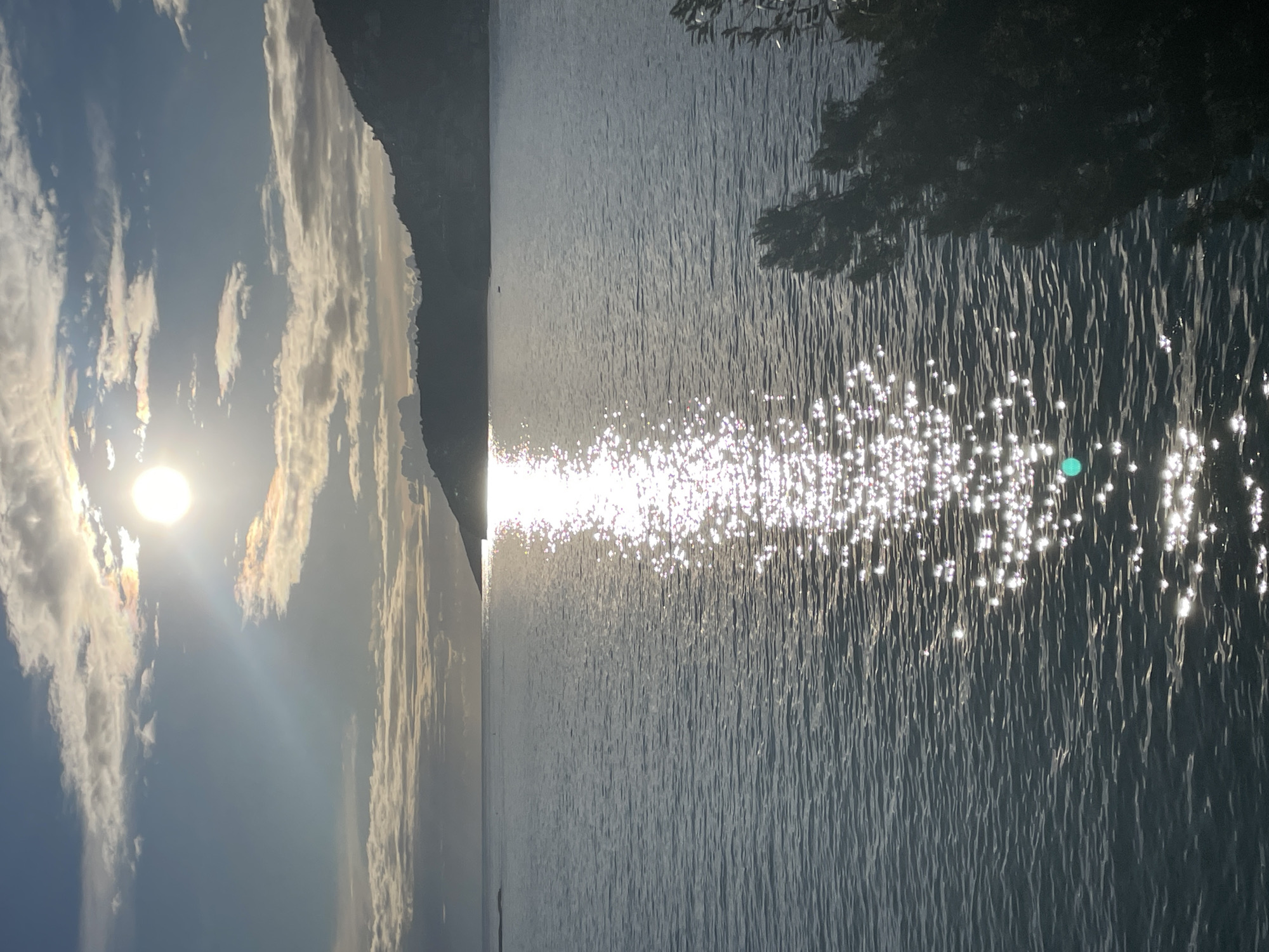 The image displays a coastal scene in Budva, Montenegro, during either sunrise or sunset. A prominent, bright, circular sun is positioned in the upper right quadrant of the sky, which contains scattered stratocumulus clouds and areas of clear blue. The sun produces an intense, vertical column of specular reflection on the surface of the Adriatic Sea, extending from the horizon towards the lower right foreground. A dark, silhouetted mountainous coastline occupies the distant horizon on the right. A small, dark, elongated object, consistent with a boat or buoy, is visible on the water's surface in the mid-ground, to the left of the sun's reflection. The lower right foreground is partially obscured by dark, silhouetted foliage. No human subjects, animals, or explicit actions are present.