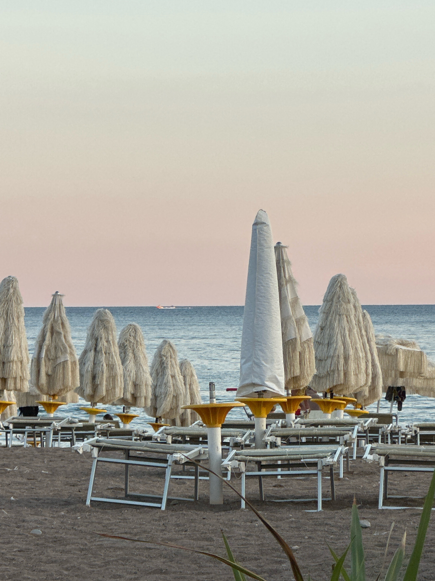 This image depicts a tranquil beach scene in Boreti, Montenegro, likely during the late afternoon or early evening. The foreground is dominated by rows of closed beach umbrellas and empty lounge chairs arranged on a sandy beach. The umbrellas vary in style; some are covered in white fabric, while others have a more natural, straw-like texture. The lounge chairs are mostly made of metal and fabric, and are neatly stacked or positioned for use.
In the middle ground, the calm, blue sea stretches towards the horizon under a soft, gradient sky that transitions from a pale blue at the top to a warm, peachy hue near the horizon. A small boat with an orange stripe is visible in the distance on the water.
The overall atmosphere is one of quietude and anticipation, suggesting the end of a beach day or the calm before it begins. No people are actively engaged in activities within the frame, contributing to the peaceful and deserted ambiance of the scene. The environment appears clean and well-maintained, characteristic of a resort or public beach area.