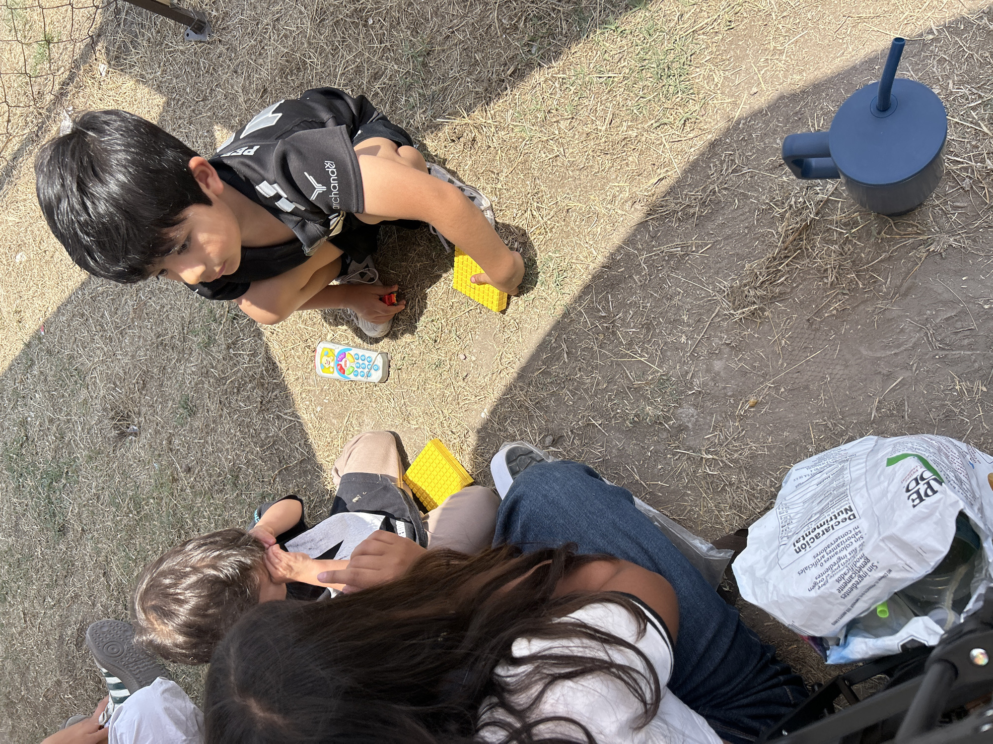 The image shows a group of people on an outdoor surface with sparse vegetation, likely grass or weeds. A young boy is crouching and playing with yellow blocks and a toy phone. Another child with long hair is sitting, interacting with the boy. An adult is visible with jeans. A blue water bottle is lying nearby. A partially visible bag labeled "PE DD" with nutritional information in Spanish is next to the adult. The ground appears to be dirt or dry soil, with some patches of vegetation. A fence is visible in the background.