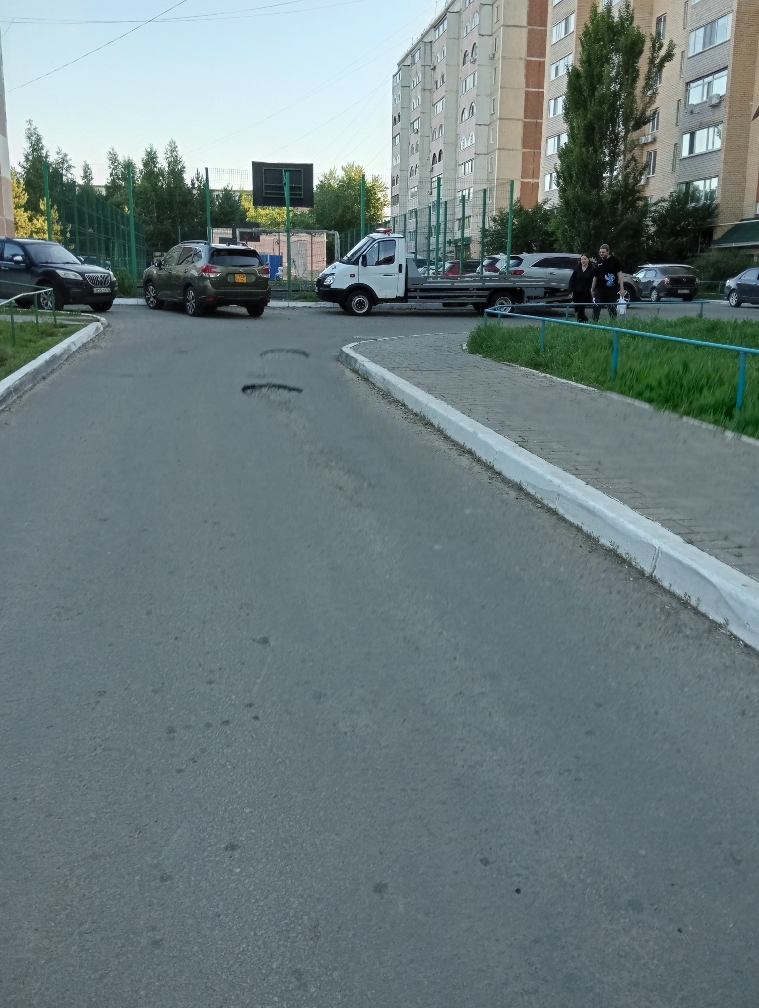 In the city of Unknown, Unknown, an outdoor scene captures a residential area with apartment buildings, a sports court, and parked vehicles. A tow truck with a white cab and a flatbed is positioned on the paved road, facing right. Two individuals, a woman and a man, walk away from the tow truck, moving from left to right. The woman is wearing a dark-colored outfit and the man is in a dark t-shirt and pants, carrying a white bag. Several cars are parked on the road, including a dark-colored SUV on the left and a green SUV in the middle. A black car is parked further down the road on the right. The environment includes a paved road with visible potholes, a sidewalk lined with grass, and a green metal fence surrounding a sports court with a backboard. The buildings are tall, multi-story apartment complexes, suggesting an urban or suburban setting. The sky is clear and light blue.