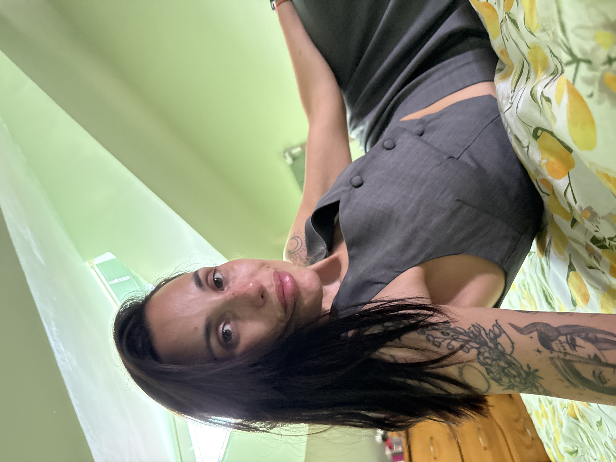 A medium shot depicts a woman with dark hair looking into the camera. She appears to be in a room with light green walls. She is wearing a gray vest-like garment. She has tattoos on her arms and upper chest. The location is unknown, with no elements to suggest Pleven, Bulgaria.