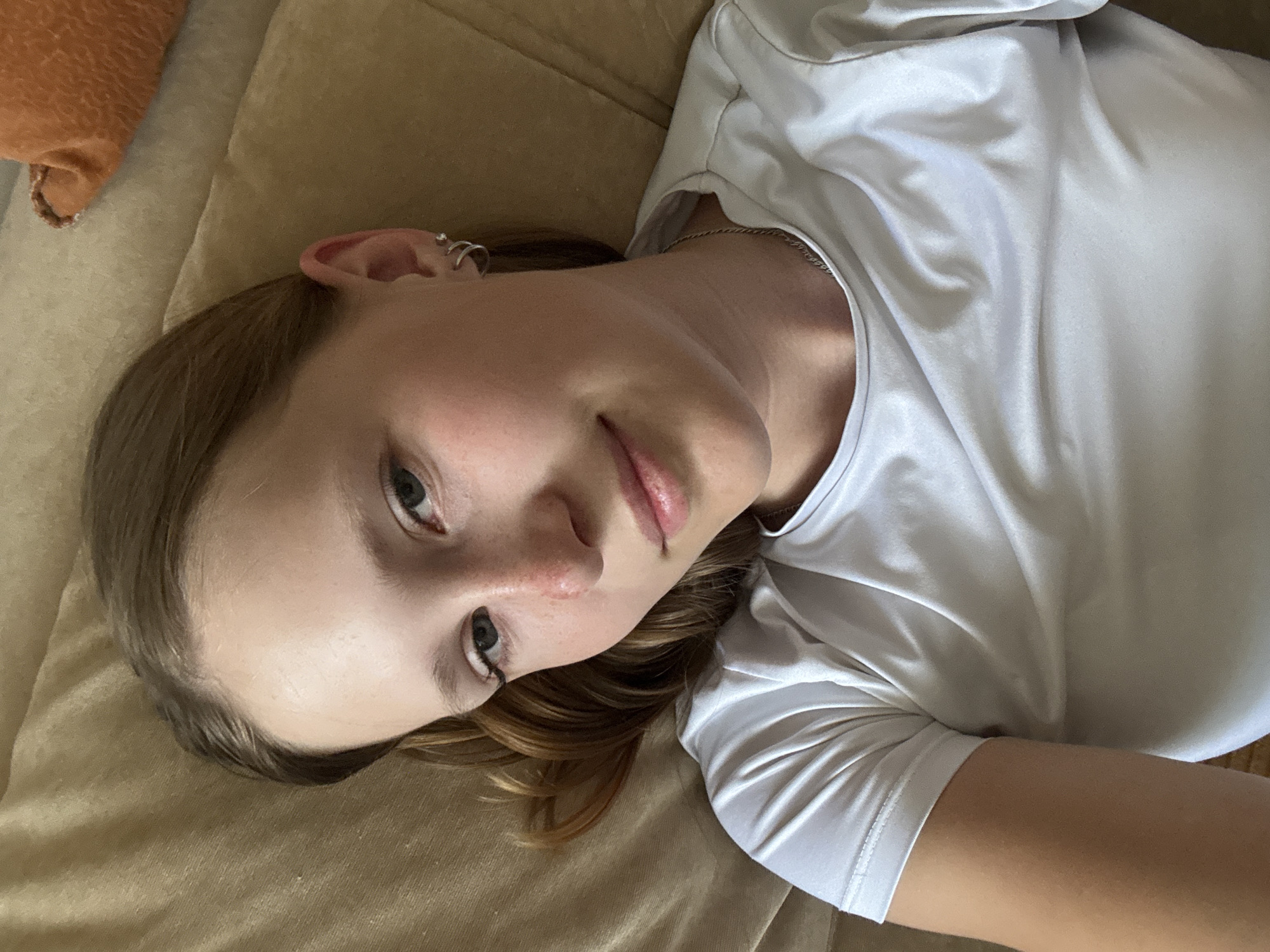 A young woman is lying down on a tan colored couch. She has light skin and light brown hair and is wearing a white shirt. She has multiple rings in her left ear. There is an orange cushion visible in the background. Location: Unknown; information about Chaikovsky, Russia cannot be determined from the image.