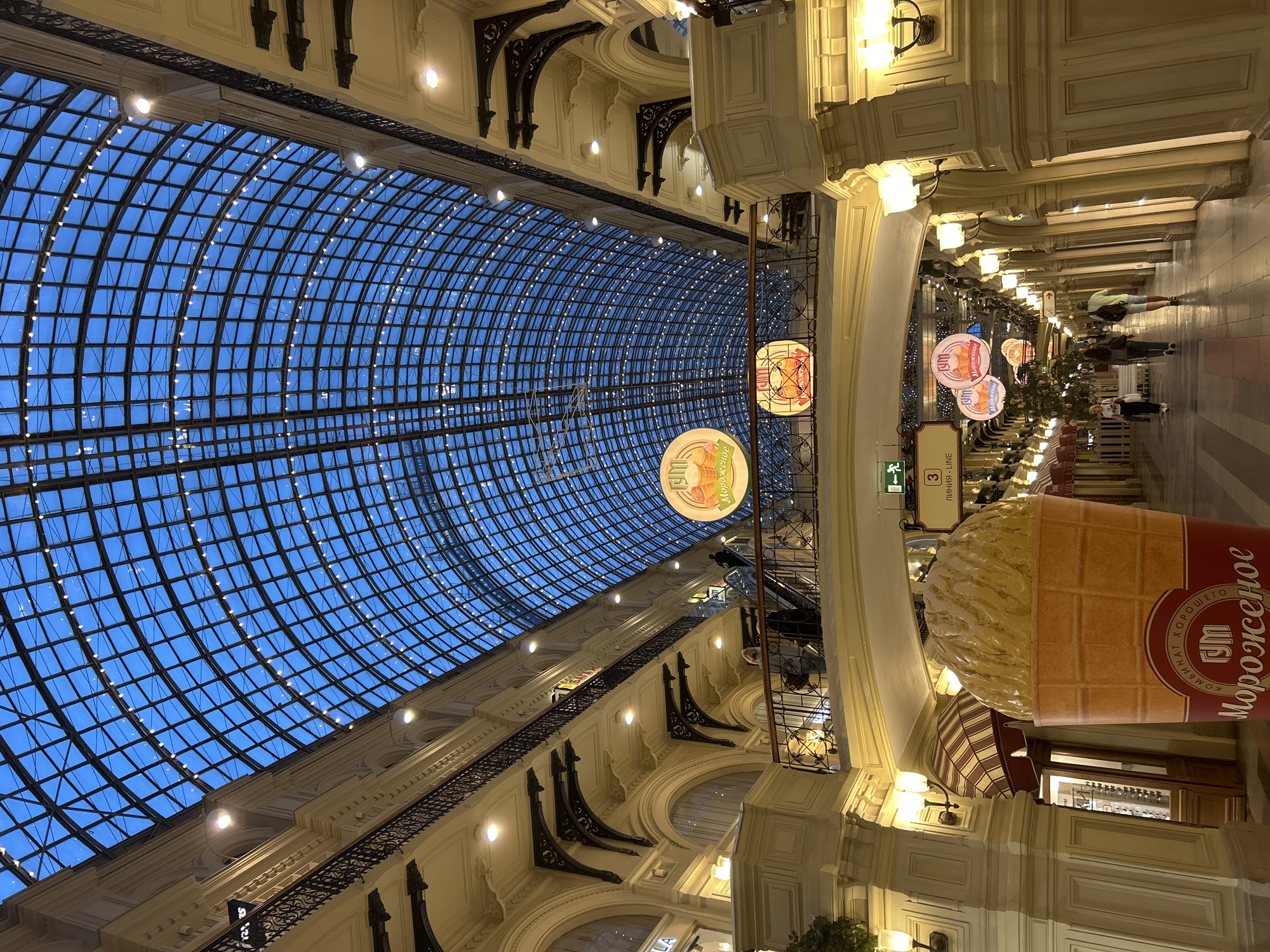 The image shows a view looking up at the arched glass ceiling of a large indoor market or department store. The ceiling is supported by a grid of metal beams, and small lights are strung along the intersections of these beams, illuminating the blue sky visible through the glass. Below the ceiling, multiple levels of walkways and balconies are visible, adorned with ornate metal railings and classical-style architecture. The walls of the building are painted a pale yellow or cream color and feature decorative cornices and arched doorways.
In the foreground, a very large, brightly lit sculpture of an ice cream cone is prominent. The cone is depicted in a waffle cone and is topped with a generous scoop of what appears to be vanilla ice cream, textured to look like soft-serve. The base of the sculpture has Cyrillic lettering on it, which translates to "GUM Ice Cream."
Further down the corridor, several people are walking, some on the ground floor and others on an upper level. Escalators and stairs are visible, facilitating movement between the levels. There are also decorative circular signs hanging from the ceiling, featuring an image of an ice cream cone and similar Cyrillic text. Lights are mounted on the walls along the corridor, casting a warm glow. The overall impression is that of a grand, well-lit indoor commercial space with a festive atmosphere, possibly during the evening or at dusk.