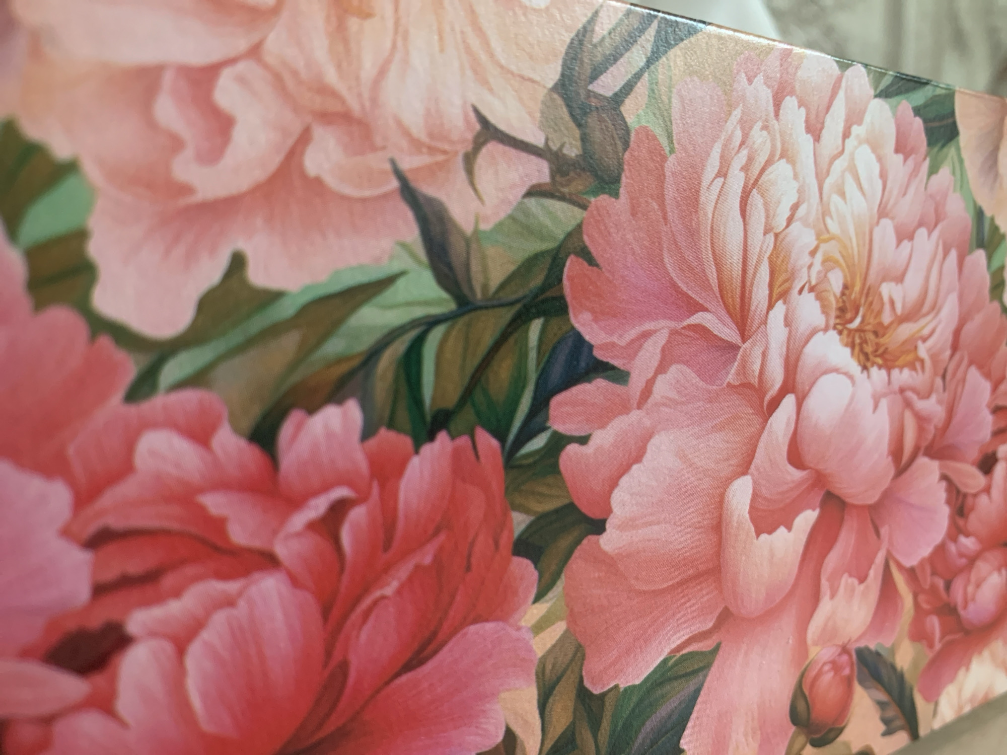 The image shows a close-up of a surface covered in a floral design featuring pink and red peonies. The leaves are shades of green and brown. The composition gives the impression of depth due to the overlapping flowers and leaves. The edge of the surface is visible in the upper right corner. There is no indication of the location being in the city of Chaikovsky, Russia.