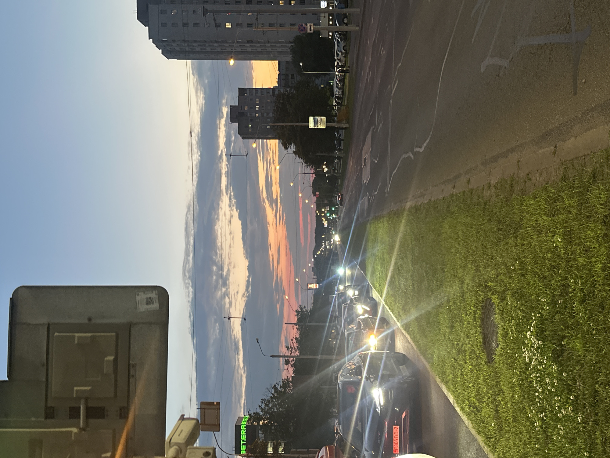The image shows a street scene at dusk in an urban environment. In the foreground on the left, there is a traffic signal pole with a camera attached. A line of cars is stopped in traffic on the left side of the road, with their headlights on. The road itself has painted lane markings and appears wet or reflective. To the right of the road, there is a patch of grass with some small white flowers and a manhole cover. In the middle ground, there are more cars and streetlights lining the road. In the background, several apartment buildings are visible, some with lights on in the windows. The sky is a gradient of colors, transitioning from blue at the top to pink and orange near the horizon, indicating sunset. There are some clouds in the sky. A sign with green lettering reading "BETARACH" is visible on a building on the left. The overall atmosphere is that of a typical evening commute in a city.