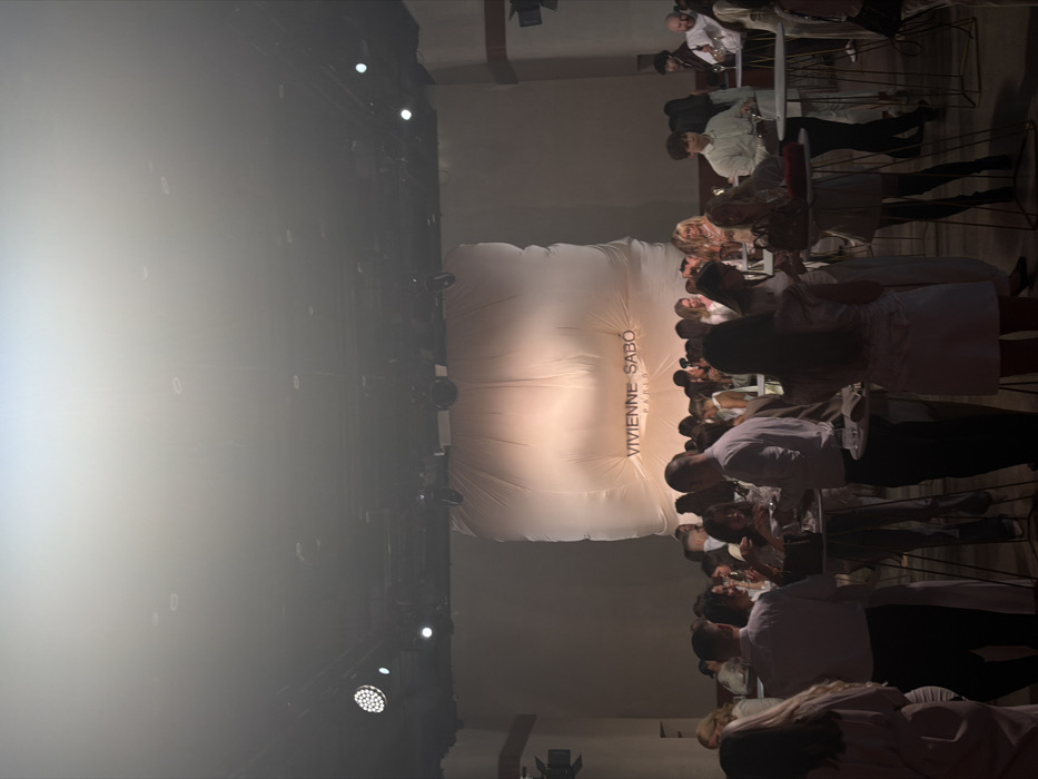 This image captures an indoor event, likely a fashion show or product launch, shot from an elevated position. The image is oriented vertically (rotated 90 degrees clockwise from its likely original orientation). If viewed correctly, the scene would show a stage or presentation area on the left and an audience seated along the right.
The left side of the frame (which would be the main stage area if correctly rotated) is dominated by a bright, diffused light source, possibly a projector or strong stage lighting, which creates a hazy, luminous effect. Several dark stage lights or spotlights with bright circular lenses are mounted on a dark truss or overhead structure, visible along the top and bottom edges of this bright area (which would be the left and right sides of the stage area). The background behind the bright light is dark and obscured by the light and haze.
In the center of the image, acting as a backdrop for the event, is a large, white, rectangular structure, possibly an inflatable screen or a fabric installation, displaying prominent text. The text reads "VIVIENNE SABO" in a dark, stylized font, with "PARIS" written in smaller letters directly below it, indicating the brand associated with the event. This backdrop has visible folds and creases, suggesting its material and construction.
To the right side of the image (representing the audience area if correctly rotated), numerous people are seated at tables arranged in rows. These tables are small, square or rectangular, with thin, dark metal frames. The seating consists of dark chairs, possibly bar stools, accommodating the attendees. The audience members, predominantly adults, are engaged in various activities: some are looking towards the left (the stage area), others are conversing, and a few appear to be holding drinks or looking at their phones. Their attire varies, with several individuals seen in light-colored shirts or dresses. The lighting in the audience area is dimmer and warmer compared to the bright, cool light on the stage, creating a focus on the central presentation space.
The overall atmosphere suggests a formal yet social gathering. The flooring on the audience side appears to be a light, possibly concrete or tiled, surface. In the far right background (which would be the ceiling or wall if correctly oriented), structural elements like beams or supports are faintly visible, contributing to an industrial or modern aesthetic for the venue.