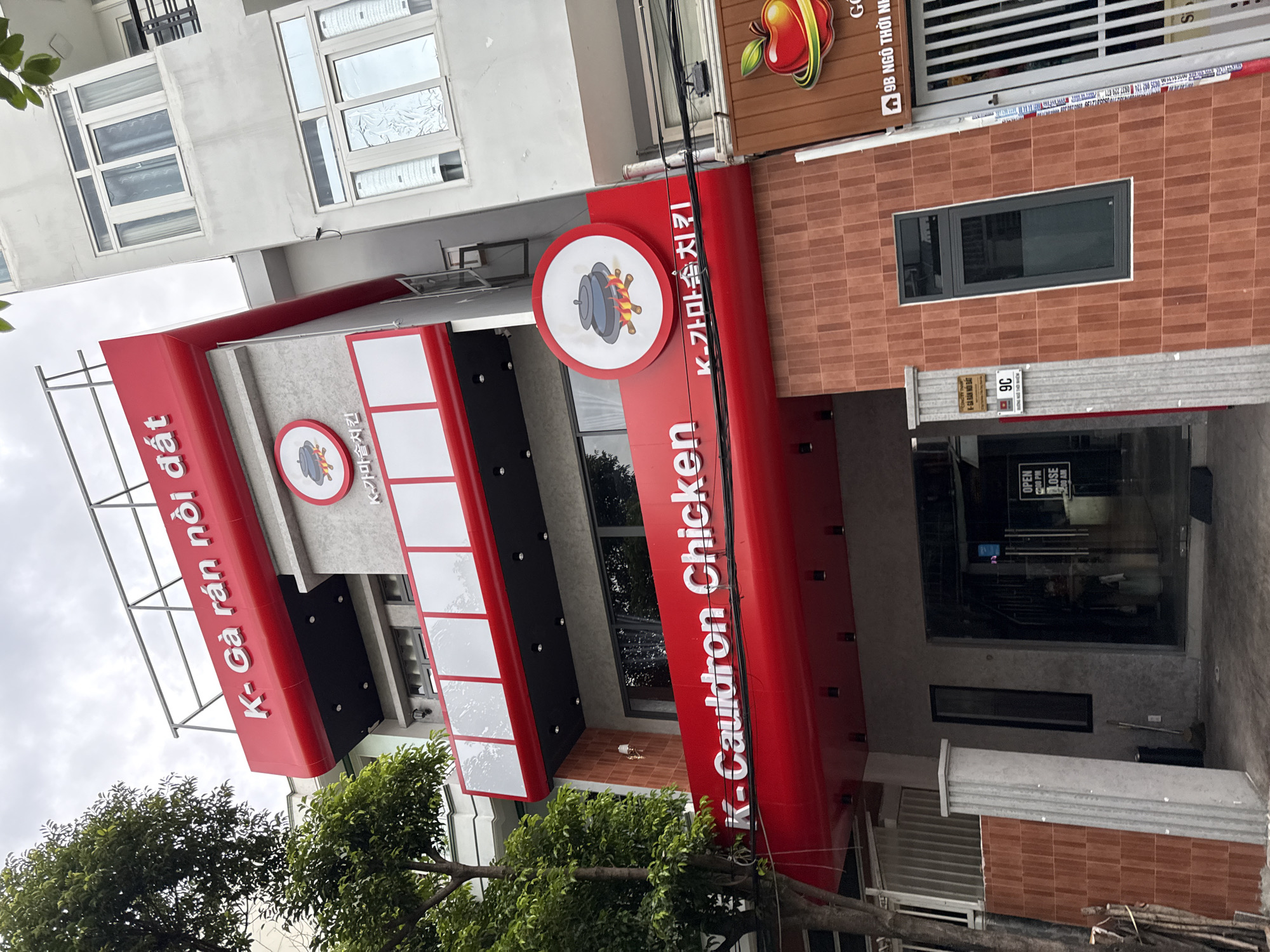 The image shows the exterior of a multi-story restaurant with a red and white facade in Nha Trang, Vietnam. The restaurant is named "K- Cauldron Chicken" and features Vietnamese and Korean text on its signage. The building has several windows and a prominent red awning. A tree with green foliage is visible on the left side of the image. A sign with an apple graphic and Korean text is also present on the right side of the building. The sky is overcast. There are no people visible in the image.