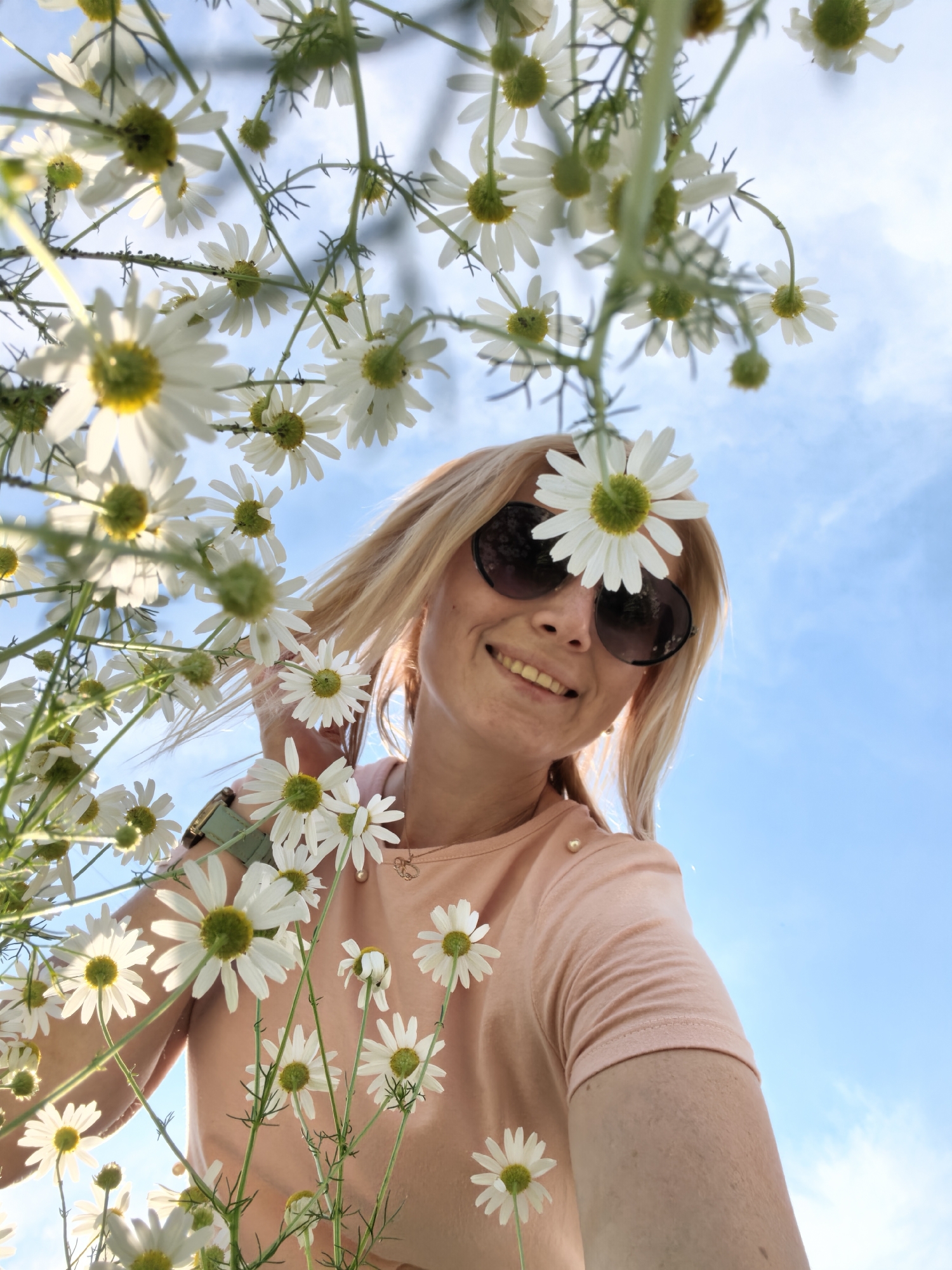 A blonde woman with sunglasses and a peach t-shirt smiles at the camera, holding a daisy above her head. Daisies are blooming all around her against a bright blue sky with some clouds. The woman appears to be in a field or park.
