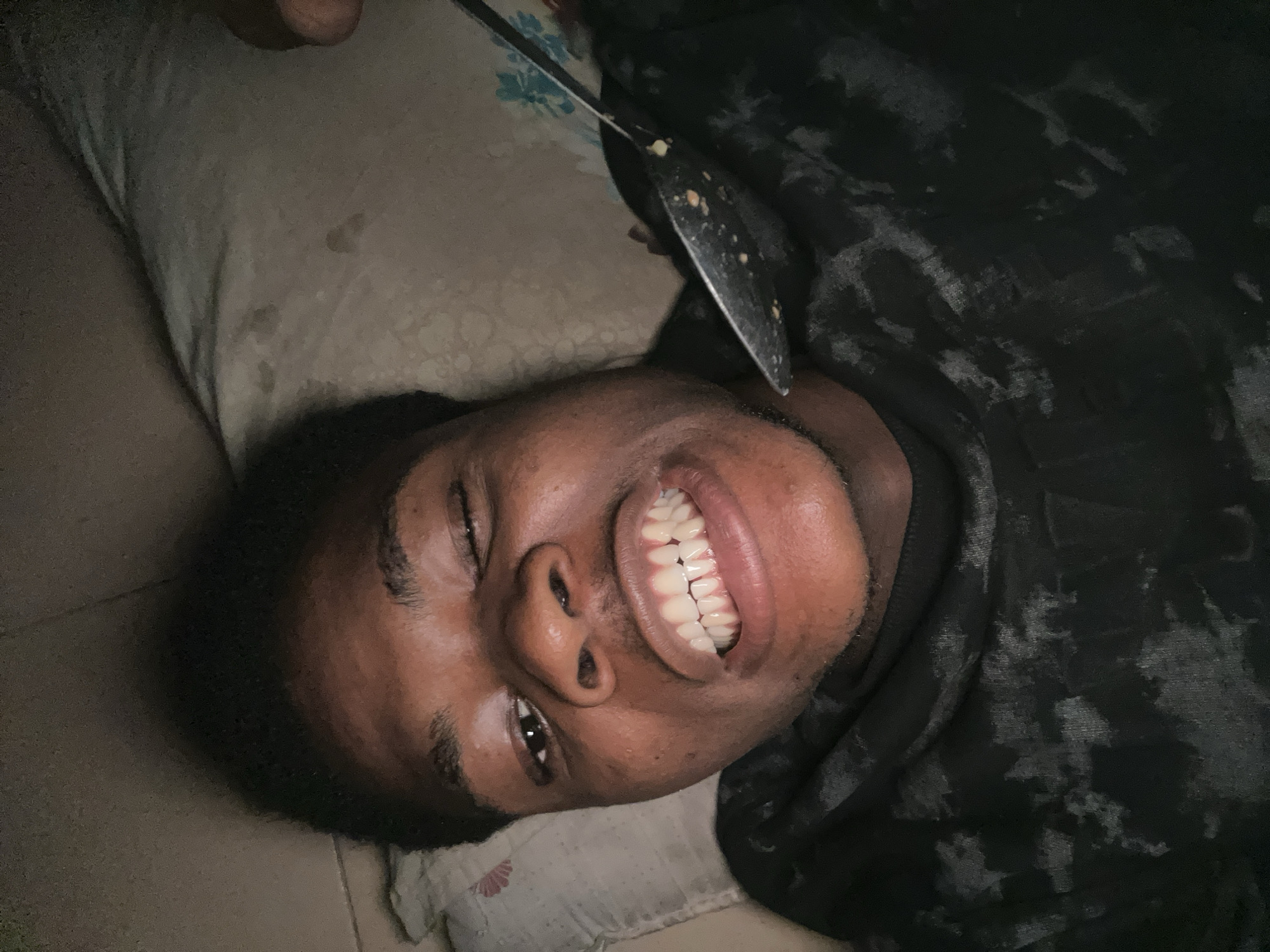 In Ihiagwa, Nigeria, a person is lying on their back on a pillow, with their head turned to the side. They are wearing a dark, patterned shirt. A metal spatula, speckled with food remnants, is positioned above their head. The person has dark skin, dark hair, and is smiling with their teeth showing. Their left eye is closed, and their right eye is open. The pillow they are lying on appears to be light-colored with some faint stains. The overall environment is dimly lit.
