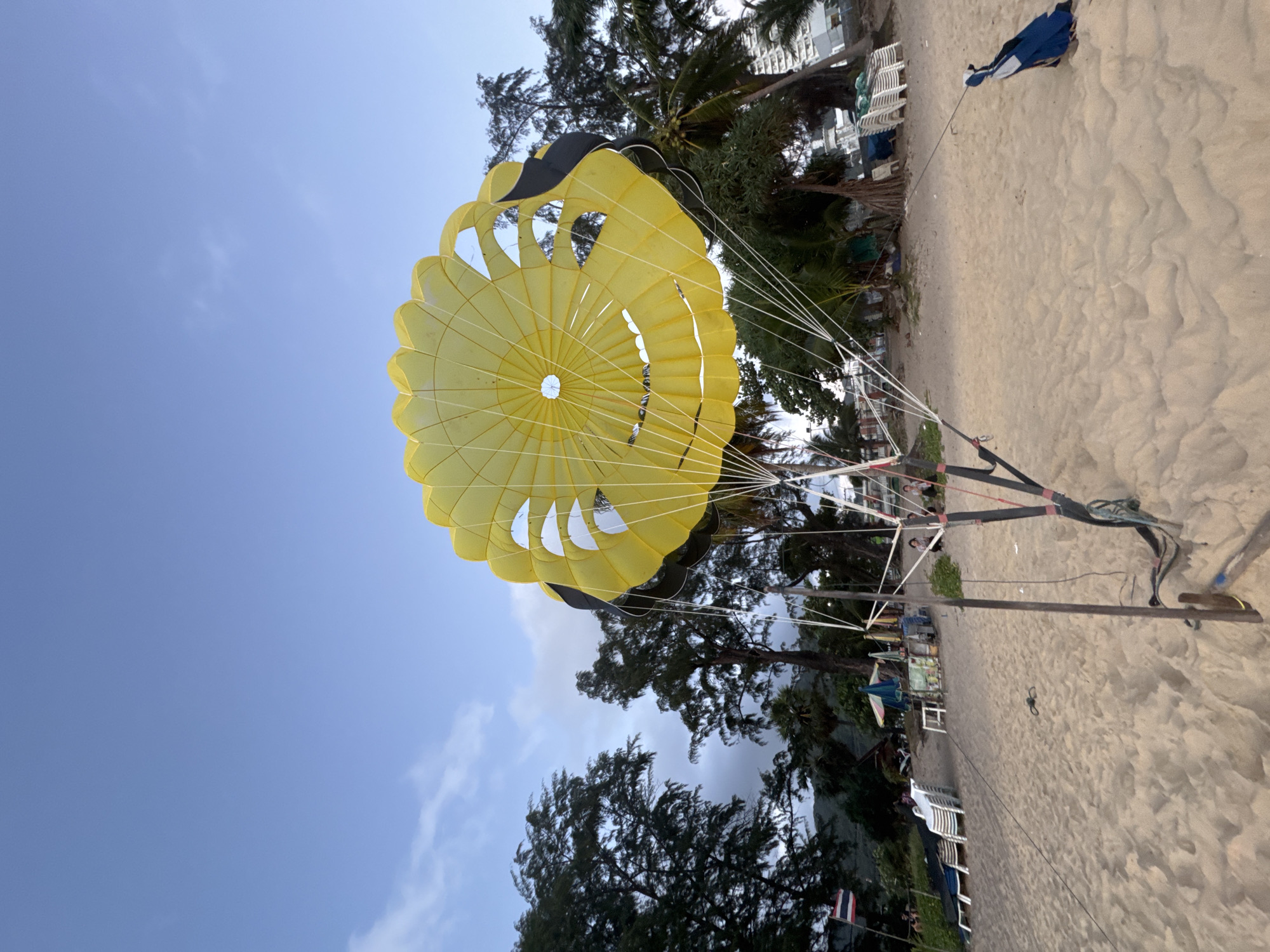 On the sandy beach of Karon, Thailand, a bright yellow parasail lies partially deployed, its canopy open to the sky. The parasail is connected to a harness system anchored to the sand by wooden posts and ropes. In the background, palm trees and lush foliage line the shore, interspersed with beach chairs, umbrellas, and small vendor stalls. The sky is a clear blue, with a few wispy clouds. The overall scene suggests a recreational area, likely a tourist destination, where parasailing activities are offered.