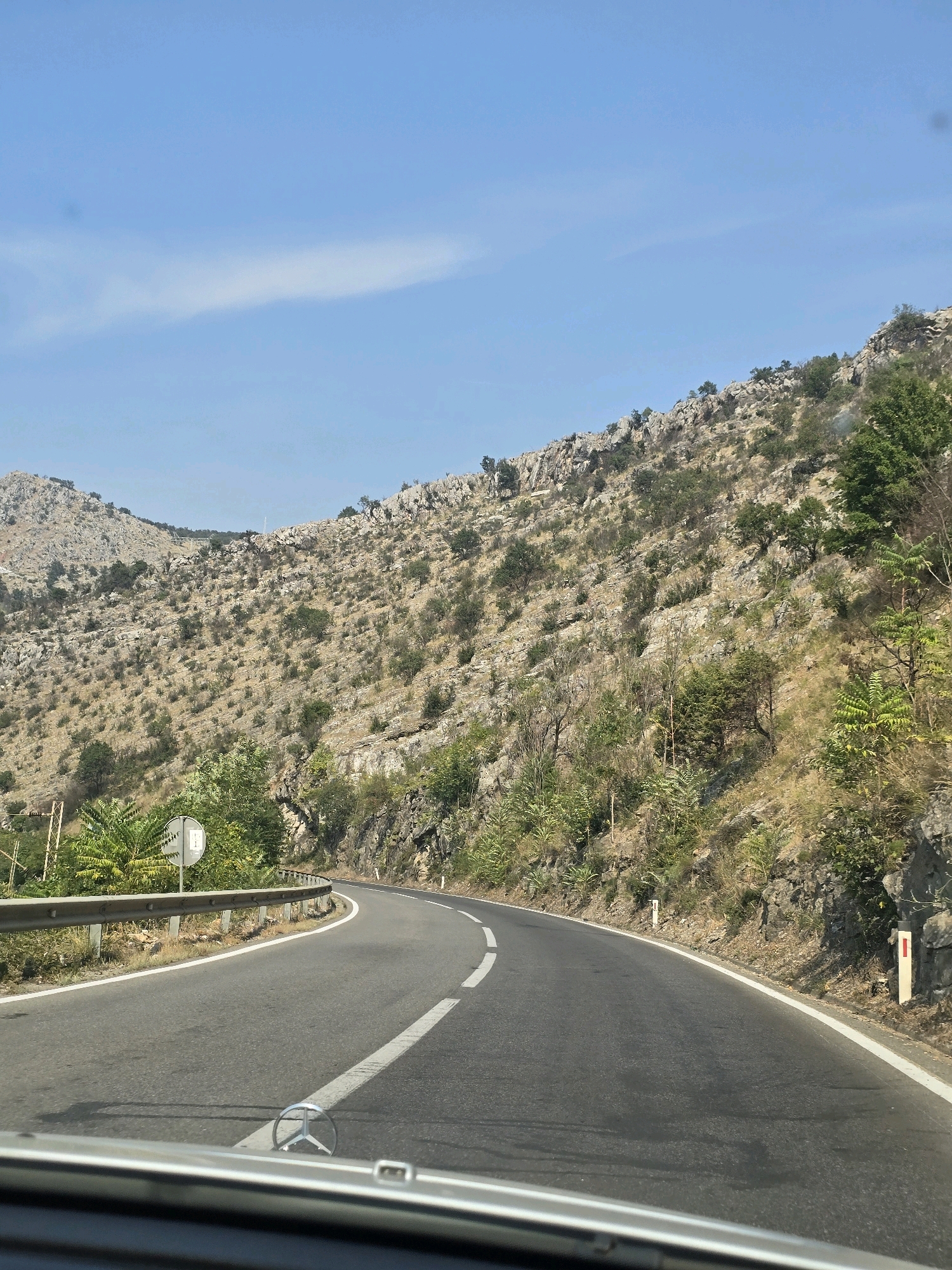 A paved road curves to the right, bordered by a metal guardrail on the left and a rocky, sparsely vegetated hillside on the right. The hillside is covered with dry grass, rocks, and scattered green trees and bushes. The sky above is clear blue with a wispy white cloud. The foreground shows the dashboard of a car, with a Mercedes-Benz emblem visible on the hood. This scene is likely a road leading out of Podgorica, Montenegro, into the mountainous region surrounding the city.