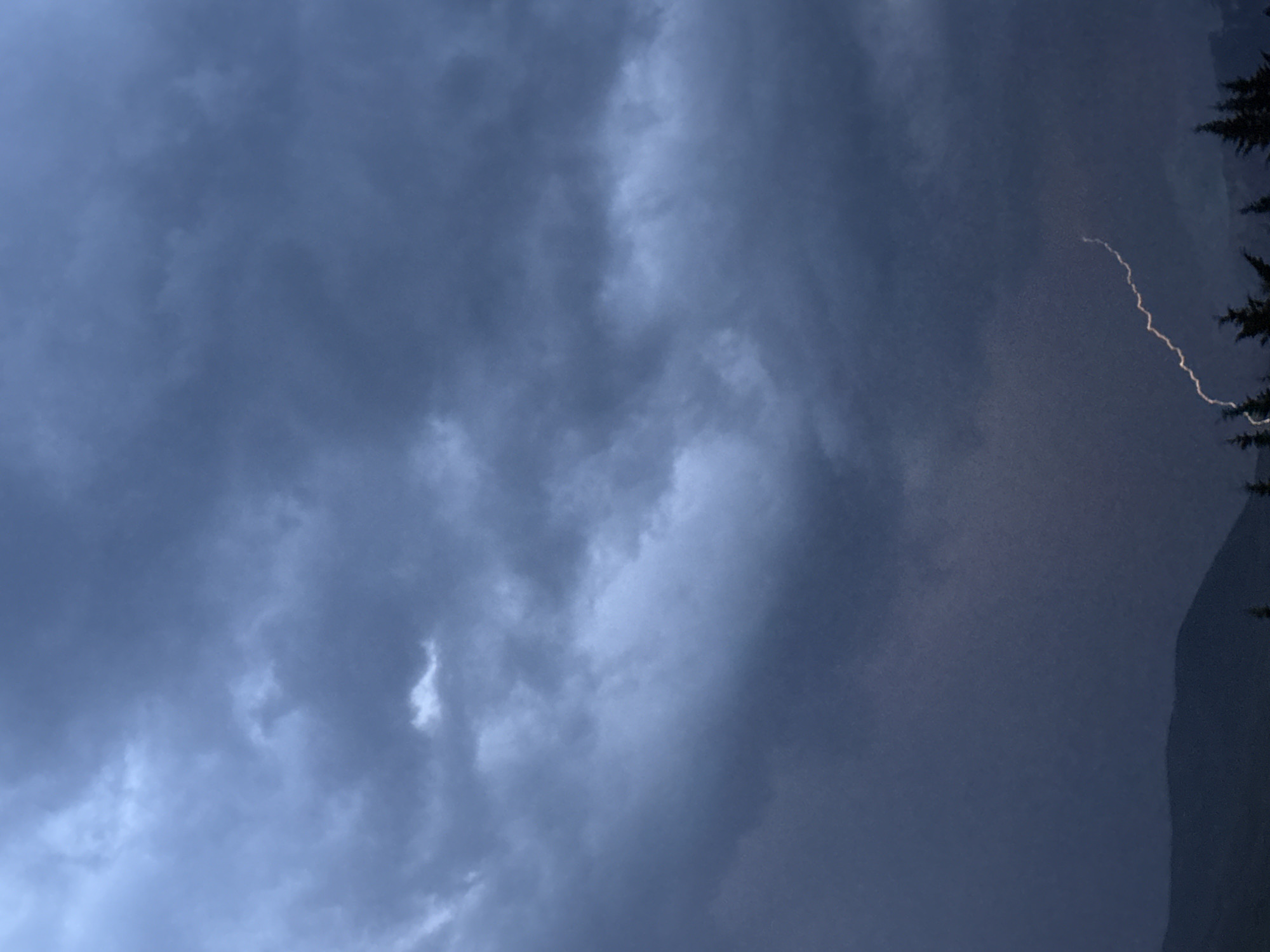 The image displays a dramatic, overcast sky dominated by dense, dark blue and gray clouds. The heavy cloud cover suggests either a late evening or early morning scene, or more likely, conditions during a thunderstorm due to the atmospheric lighting. In the upper right quadrant, a bright, jagged line, characteristic of a lightning strike, cuts across the darker sky. To the far right, partially visible silhouettes of evergreen trees rise against a dark, indistinct background, which appears to be a mountain or hillside. The perspective is looking upwards, capturing the sky and the top edges of the landscape. No people, other specific objects, or discernible text are visible. The overall mood is powerful and potentially awe-inspiring, indicative of volatile weather. The mountain and tree silhouettes align with the provided location context of Benasque, Spain.