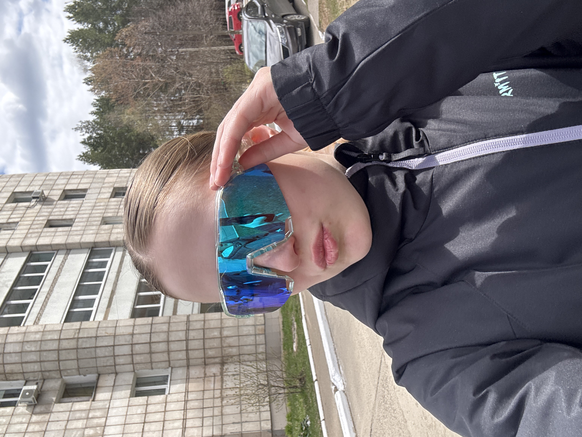 A person wearing oversized iridescent sunglasses is holding their hand up to the glasses in an outdoor setting. They are wearing a black jacket with a light-colored zipper. A light-colored building with many windows is in the background to the left. Trees and cars are visible further in the background. The scene appears to be in Chaikovsky, Russia, based on the architecture of the building.