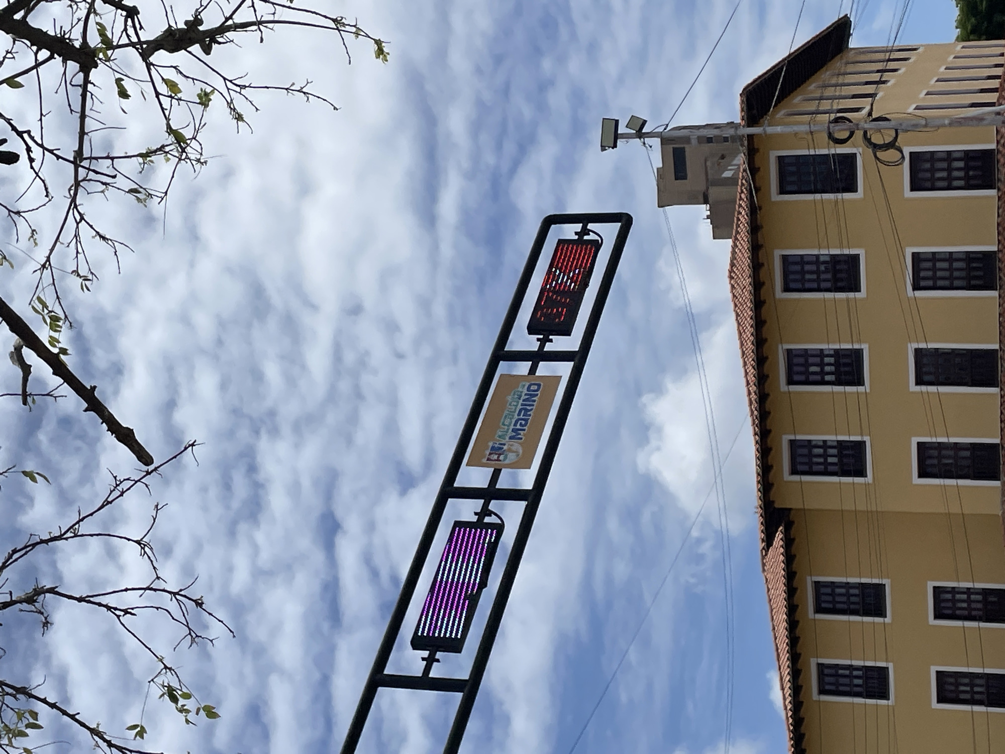 The image captures a street-level view in Porlamar, Venezuela, focusing on a traffic light and a building. A black metal frame, suspended overhead, holds two illuminated displays. The left display shows rows of pink and white horizontal lines, while the right display shows bright red text or symbols. To the right of the displays, a rectangular yellow sign with a logo and the text "ALCALDÍA MARINO" is visible. In the background, a light yellow building with a red tile roof and numerous dark-colored windows stands prominently. Above and behind the yellow building, a taller, light brown building can be seen. The sky is filled with white, fluffy clouds against a blue backdrop. Branches of a tree are visible in the upper portion of the image.