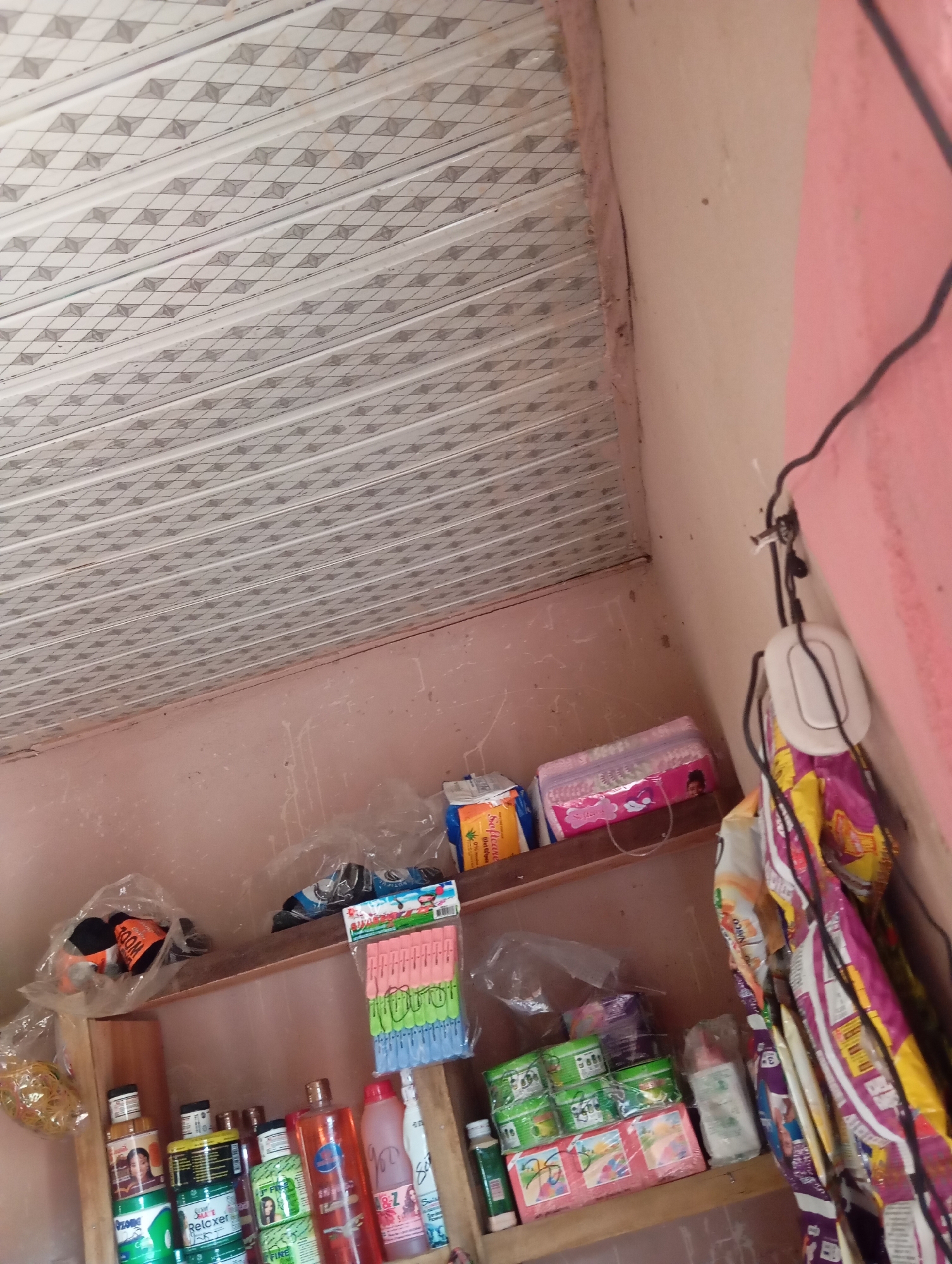 The image shows an indoor scene, likely a small shop or market stall, located in the city of Jalingo, Nigeria. The room is dimly lit and appears cluttered with various goods. In the foreground, shelves are stocked with an assortment of products, including bottles of liquid, likely toiletries or cleaning supplies, some with labels featuring images of people and text in English and possibly other languages. There are also packages of what appear to be feminine hygiene products, snacks, and bags of rubber bands. The ceiling is visible at the top of the image, with a patterned design. A bundle of clothes pegs in various colors is also displayed on a shelf. The walls are painted a light pink color. The overall impression is of a retail space with a wide variety of items for sale, typical of a small business in Nigeria.