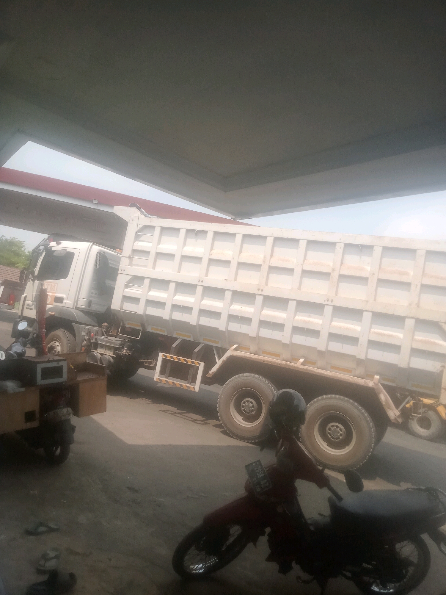A white dump truck is parked under a canopy, possibly a gas station or a covered parking area. The truck's bed is raised. In the foreground, a red motorcycle is parked, and its rider, wearing a helmet, is seated on it. The ground appears to be asphalt. The scene is outdoors, with a cloudy sky visible above the canopy. The location is identified as Lebaksiu, Indonesia.