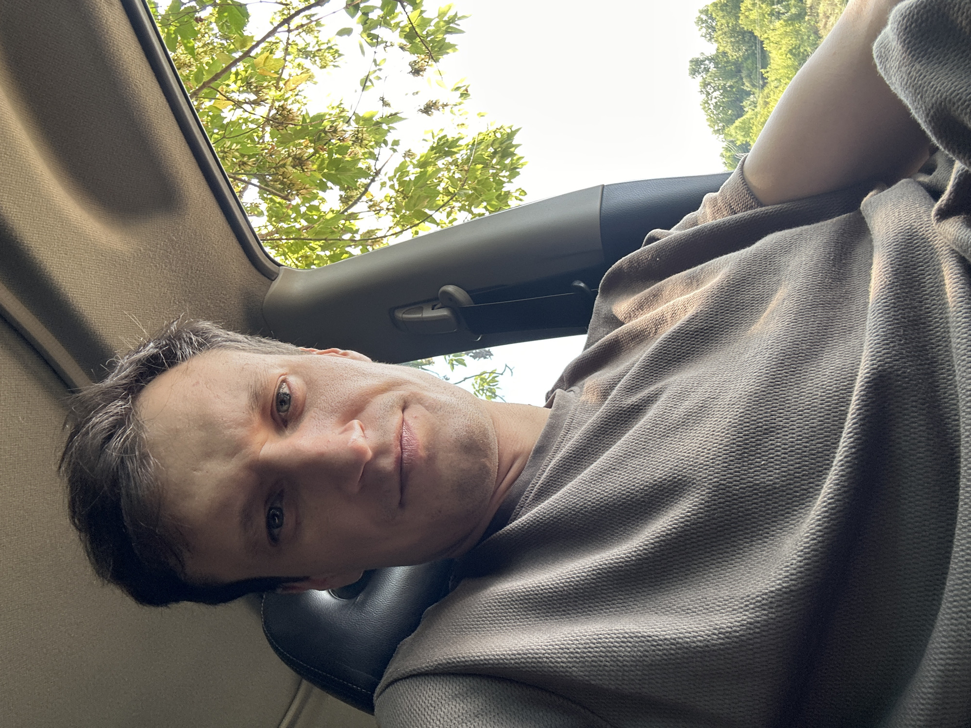 A selfie taken from inside a vehicle shows a man with short, dark hair and light skin. He is wearing a brown, textured long-sleeved shirt. The man is looking directly at the camera with a neutral expression. To his right, a car's interior is visible, including a dark seat and a grey ceiling. Outside the car window, sunlight filters through green leaves of trees, and a bright sky is visible. The overall environment suggests a daytime scene, possibly during a drive. The exact location in Makiv, Ukraine, cannot be determined from the image alone.