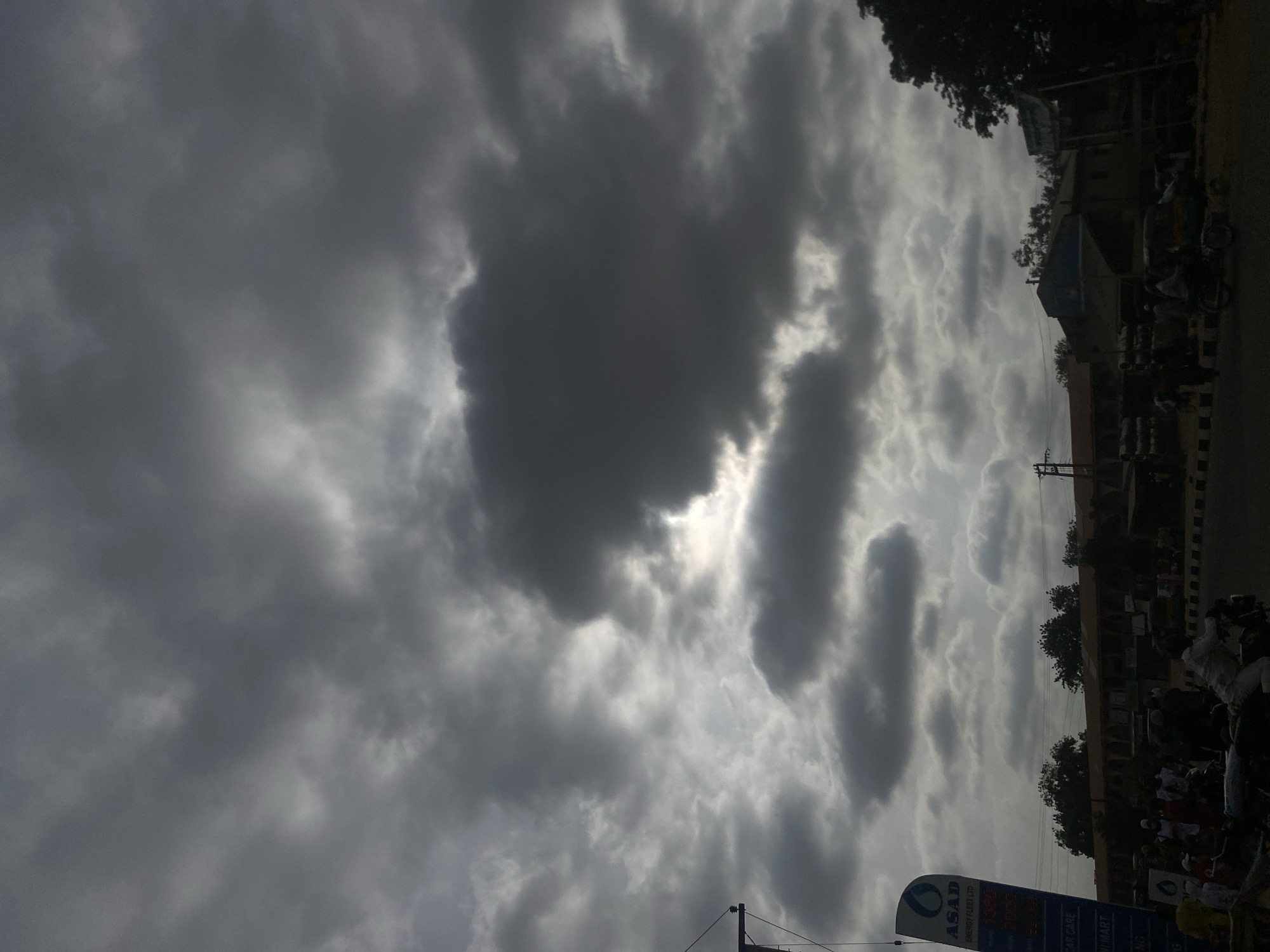 The image shows a cloudy sky over Tudun Wada, Nigeria. The sky is dominated by dark, heavy clouds, obscuring much of the sunlight. Below the clouds, a street scene is visible. Buildings line the street, with trees interspersed among them. There are parked motorcycles, and people are visible on the sidewalk. An advertising sign with "ASAD" written on it is in the foreground.
