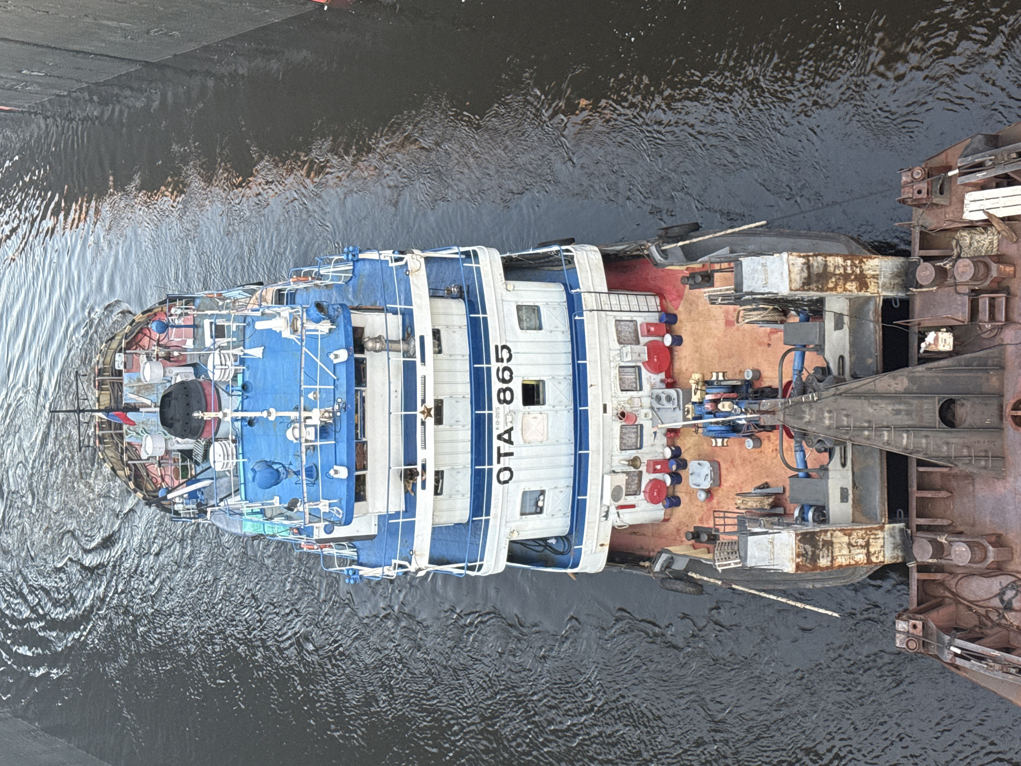 An aerial, eye-level shot shows two connected barges traveling on a dark, choppy body of water. The barge in the foreground is rusty and brown, with a prominent black, pointed structure at its front. It is connected to a larger, white and blue barge behind it. The second barge has multiple decks, a white cabin with blue trim, and a blue roof. The number "OTA 865" is visible in black lettering on the side of the white cabin. The deck of the second barge is painted red and is equipped with various pieces of equipment, including winches, ropes, and several red and blue barrels. A Russian flag is visible on the top deck of the second barge. The water surrounding the barges is dark and appears to be a canal or a river, with ripples and waves creating a textured surface. The sides of the waterway are lined with dark, vertical structures that suggest walls or embankments. The scene is captured from a high vantage point, looking down onto the barges and the water.