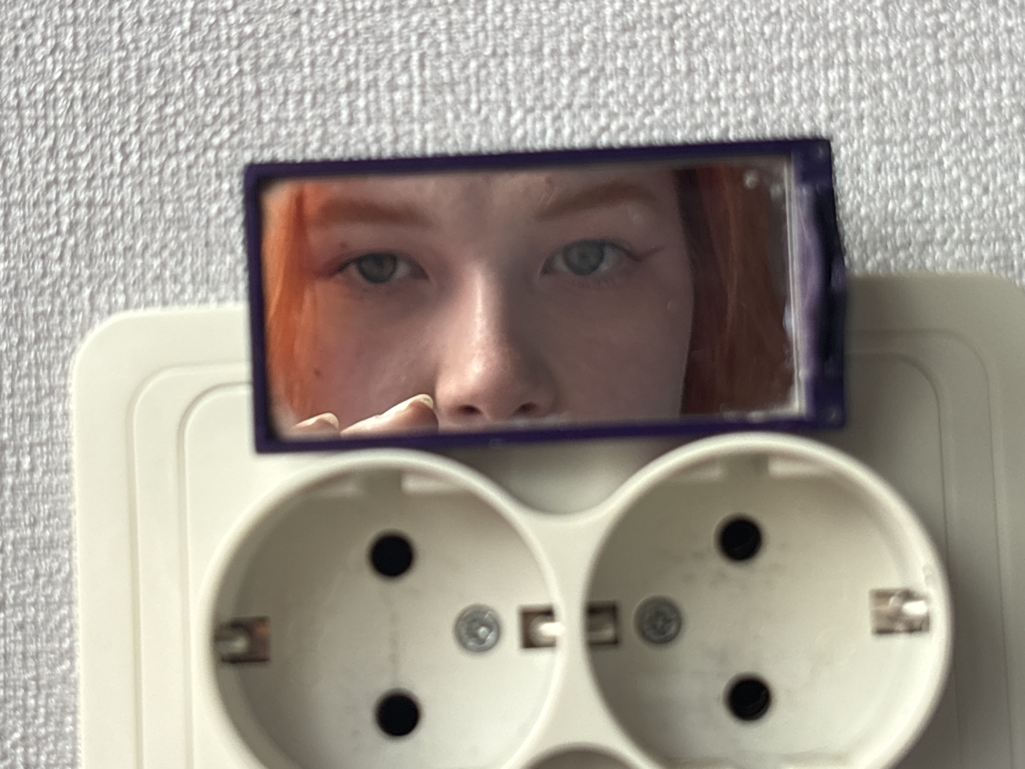 The image shows a white, European-style electrical outlet with two sockets mounted on a wall with a textured, off-white surface. A small, rectangular mirror with a dark purple frame is positioned on top of the outlet. The reflection in the mirror shows the face of a fair-skinned person with red hair, focusing on their eyes, nose, and part of their forehead. A finger is visible in the lower left corner of the mirror, possibly indicating the person is holding it. The location in the city of Unknown, Unknown is not specified.