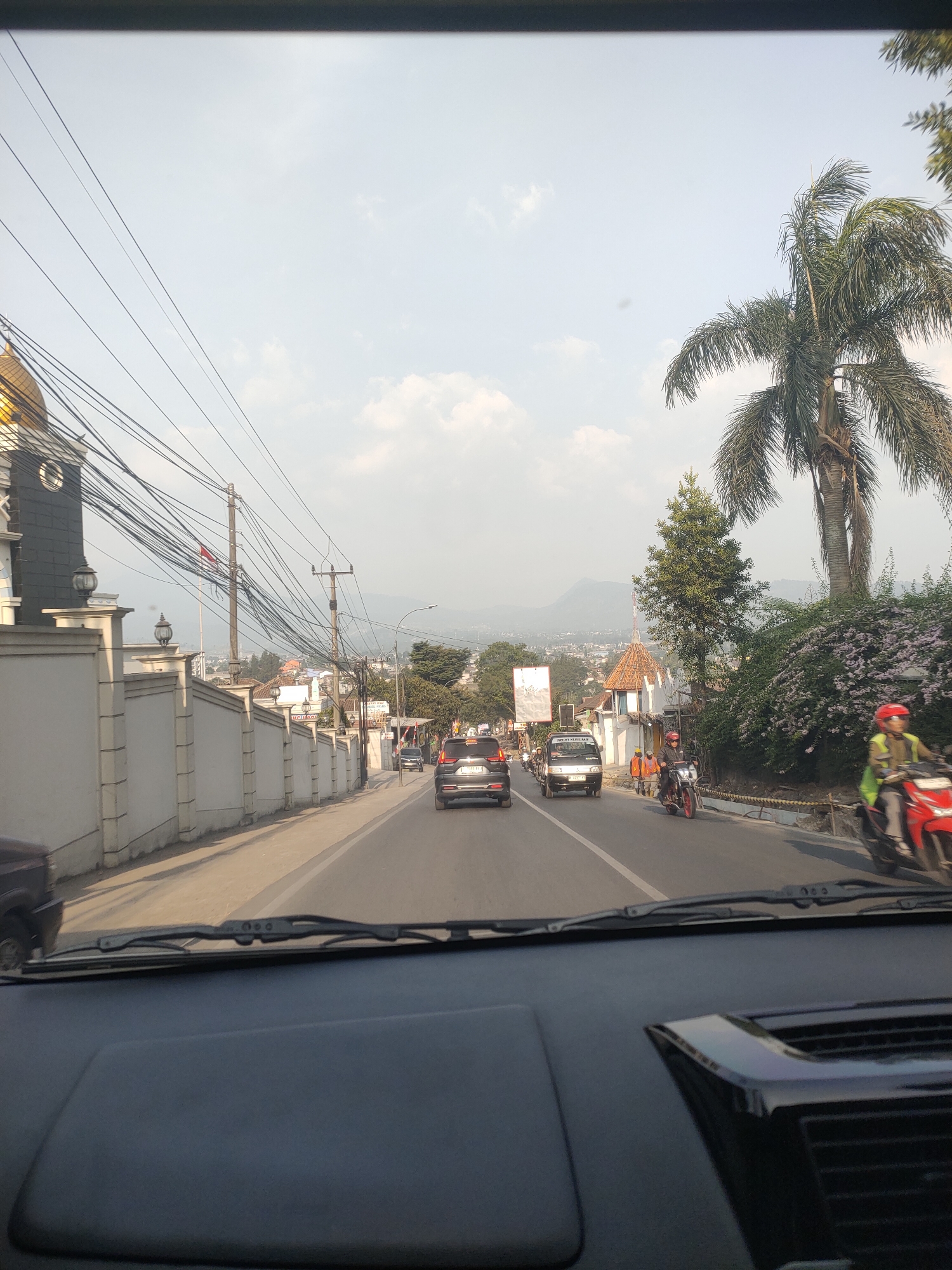 The image depicts a street scene from inside a vehicle, likely in Cibogo, Indonesia. The perspective is from the driver's or passenger's seat, looking forward through the windshield. A dark dashboard occupies the foreground. The street is paved and appears to be a two-lane road with traffic.

On the left side of the road, there is a white wall with decorative elements and lamps. Behind the wall, a building with a golden dome and dark facade is partially visible, suggesting it might be a religious structure. Numerous overhead electrical wires crisscross the sky, supported by utility poles.

The right side of the road features a lush green environment with trees, including a prominent palm tree, and bushes with pink flowers. Several vehicles are on the road. In the middle ground, there are two cars, a black SUV and a silver sedan. Further ahead, a white van and a red motorcycle are visible. A person wearing a yellow vest and a red helmet is riding a red motorcycle. Another person in an orange vest is standing near the side of the road. In the background, buildings and hills can be seen, indicating a suburban or semi-rural setting. The sky is bright with scattered clouds.