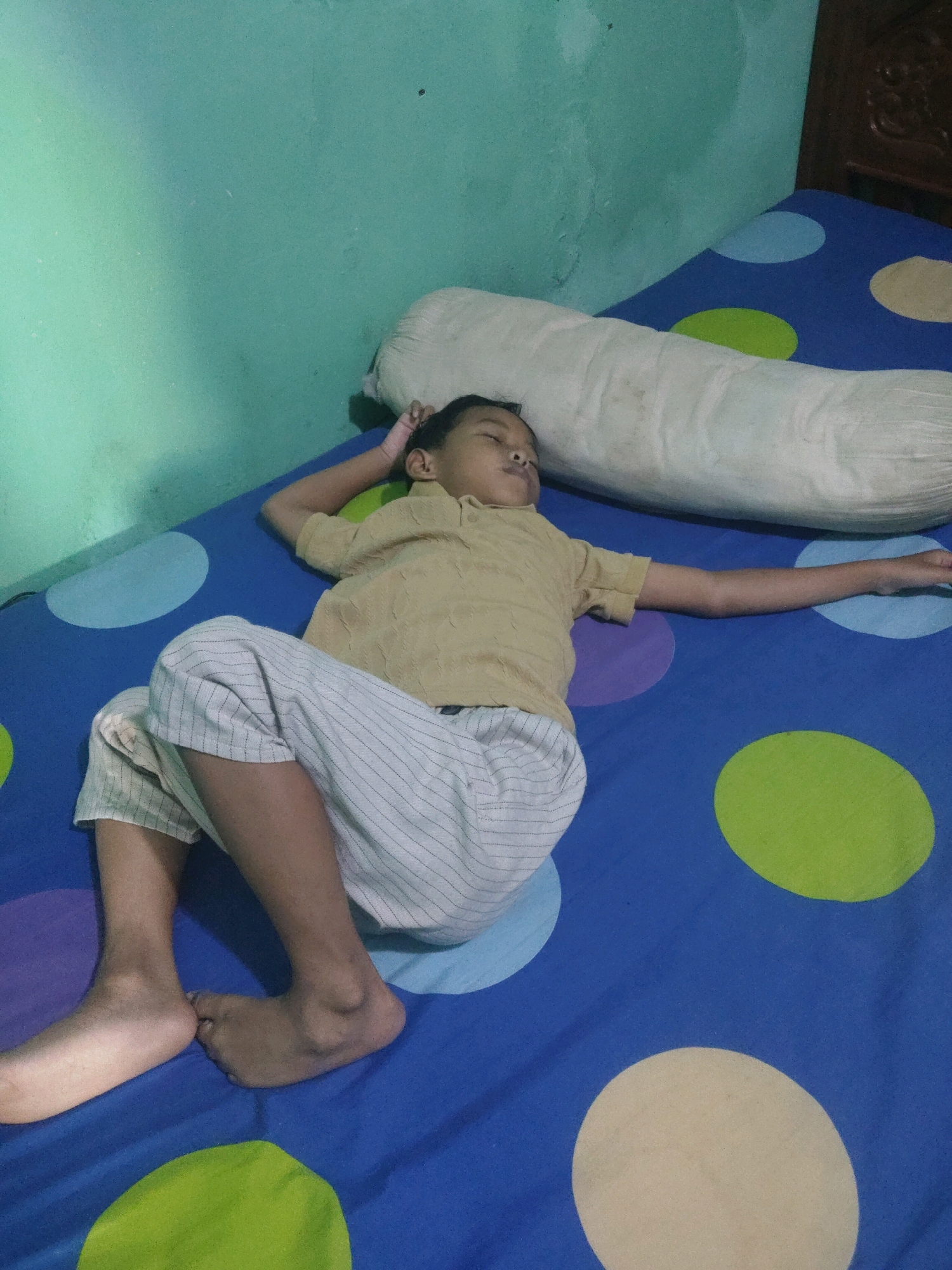 A young boy is sleeping on a bed in Subang, Indonesia. He is lying on his back with his right arm stretched out and his left arm bent. He is wearing a yellow polo shirt and white striped pants. The bed is covered with a blue sheet with large polka dots in various colors, including blue, green, and beige. A long, white bolster pillow is behind his head. The room has a light green wall.