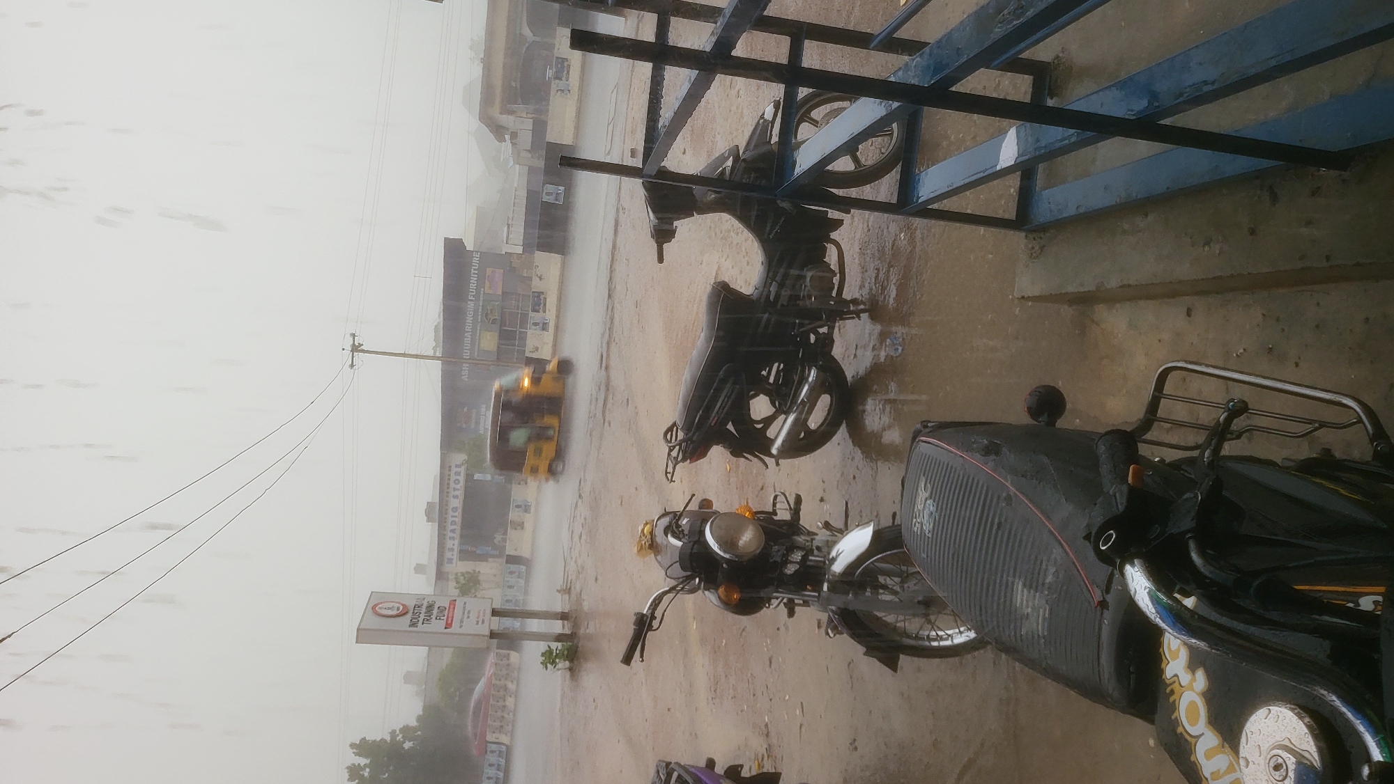 The photograph depicts a street scene in Kawaji, Nigeria during heavy rainfall. Several motorcycles are parked on a wet concrete surface, possibly under a makeshift shelter. In the background, blurred due to the rain, are buildings and a yellow motorized tricycle (keke). A sign for the Industrial Training Fund is visible. Power lines run along the street. The scene is overcast and wet.