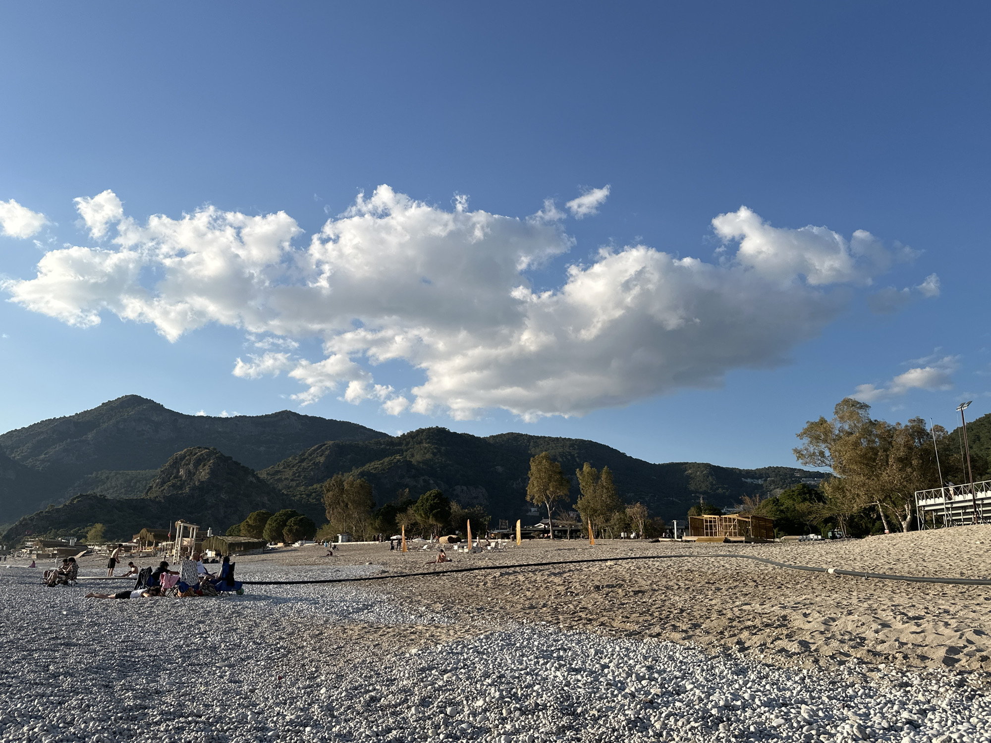 The image shows a beach scene in Fethiye, Turkey, under a partly cloudy, blue sky. The foreground features a pebble-covered area transitioning into a sandy beach. Several groups of people are sitting and reclining near the pebble section, possibly relaxing or socializing.
A dark-colored pipeline runs across the boundary between the pebbles and the sand. In the mid-ground, there are sparse trees and various structures or buildings. A mountain range forms the backdrop, completing the scene.