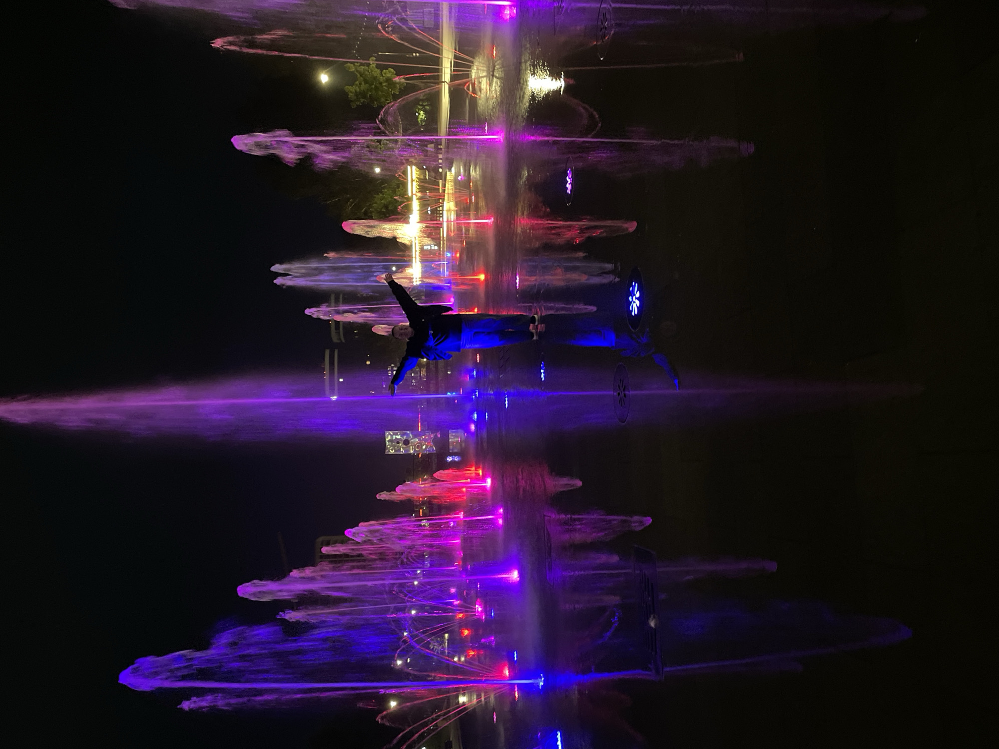 A nighttime scene shows a fountain display in Izhevsk, Russia. The fountains are illuminated with colorful lights in shades of purple, blue, and red. A person dressed in a black jacket and pants is standing in the middle of the fountain display. The surface of the fountain area reflects the lights and fountains, creating a mirrored effect. The background is dark, with some trees and buildings visible in the distance.