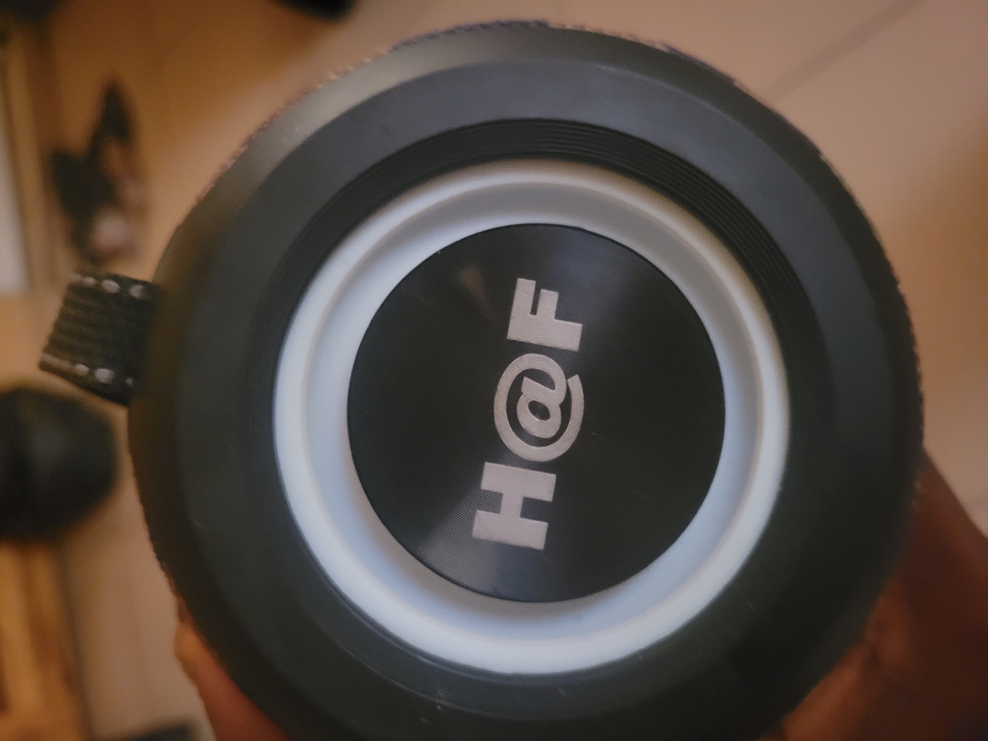 A close-up, eye-level shot shows a black portable speaker with a white ring around a black circular center. The center of the speaker has the silver logo "H@F". The speaker has a textured black strap at the top, partially visible. The background is blurred and shows a wooden surface with shadows, suggesting an indoor environment.