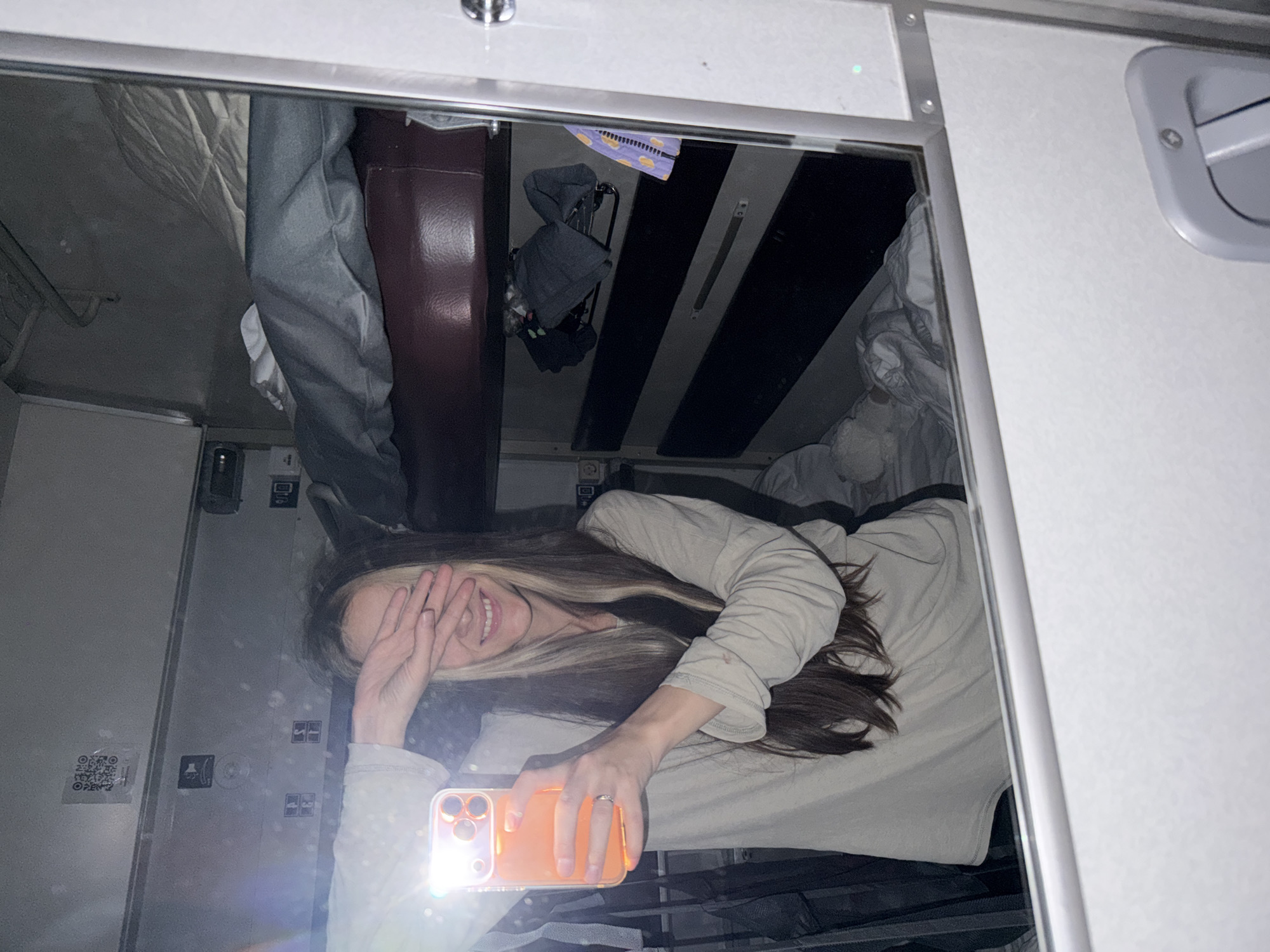 A young woman is captured in a mirror selfie, reflecting a playful moment inside what appears to be a train compartment or sleeper car. She has long, dark hair with lighter highlights, and is wearing a light-colored long-sleeved top. Her hands are partially covering her face, but a broad, joyful smile is visible. She holds an orange-cased smartphone, whose bright flash illuminates the scene and creates a lens flare. A ring is visible on her right hand.
The reflection reveals the confined interior of the compartment. Visible elements include dark striped panels on the ceiling, a dark red/brown upholstered surface (likely a seat or bunk), and light-colored bedding. Hanging near the ceiling is a dark, folded item, possibly clothing on a hanger, and a purple item adorned with yellow dots. The light grey/white walls are equipped with various fixtures, buttons, and a small grey handle or latch on what could be a door or storage compartment.
A QR code sticker with the word "OPEN" visible below it is present on the reflected left wall. The use of the phone's flash suggests a dimly lit environment, possibly indicating nighttime. The functional, compact nature of the space, along with elements like bunk beds and specific fixtures, strongly indicates a train cabin, consistent with a travel setting in Kirsanov, Russia. The woman's pose and smile convey happiness and a relaxed mood.