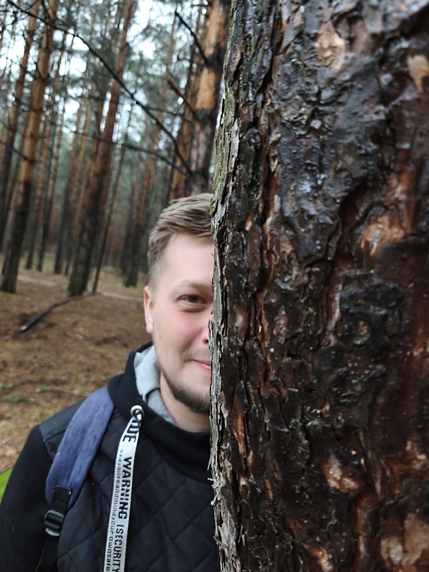 A medium close-up shot captures a man partially concealed behind a large, dark tree trunk in a pine forest, likely in Chaikovsky, Russia. The man's right eye, nose, and mouth, revealing a slight, pleasant smile, are visible as he peeks from behind the tree. He has short, light brown hair and a light beard. He wears a dark, possibly quilted jacket over a grey hoodie, and a blue backpack strap is visible on his left shoulder. A white lanyard hangs around his neck, displaying text.
The tree trunk in the foreground is prominent, with rugged, dark bark that appears wet, suggesting damp weather or recent rain. In the background, numerous tall, slender pine trees create a dense forest canopy, blurred to emphasize the man and foreground tree. A path or clearing is visible on the forest floor, which is covered with fallen leaves or pine needles. The lighting is soft and diffused, indicative of an overcast day. The visible text on the white lanyard reads: "WARNING !SECURITY savavliveandmindyour ownsafe". The overall scene suggests a candid and slightly playful moment in a natural, serene setting.