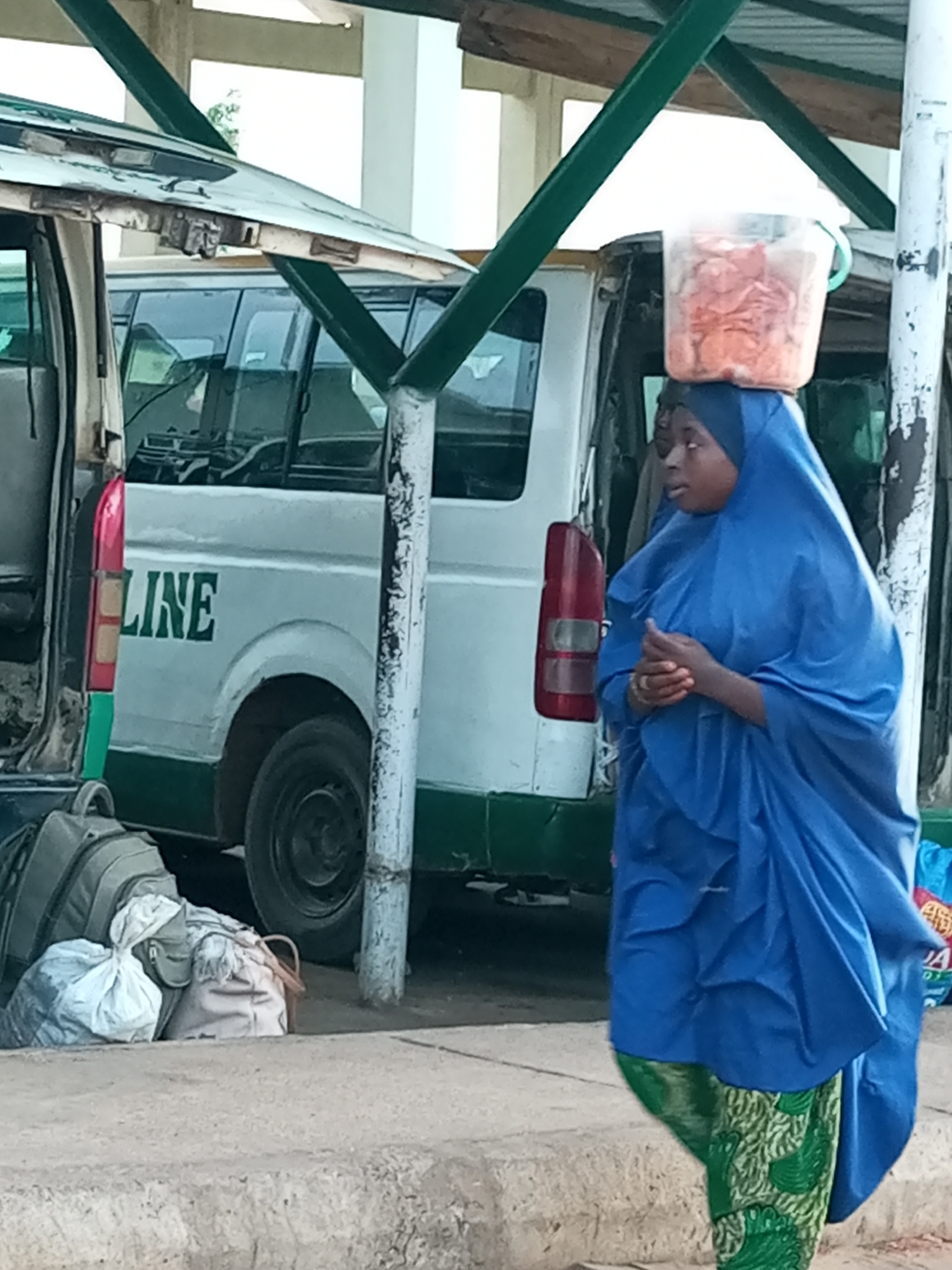 A woman wearing a blue hijab and a patterned green skirt carries a clear plastic bucket filled with orange snacks on her head. She is walking beside a white van with "LINE" written in green lettering on its side, which appears to be a public transport vehicle. Several bags are stacked on the ground next to the van. The scene suggests a bustling transport hub or market area in Gombe, Nigeria, possibly indicating the woman is a vendor or a traveler.
