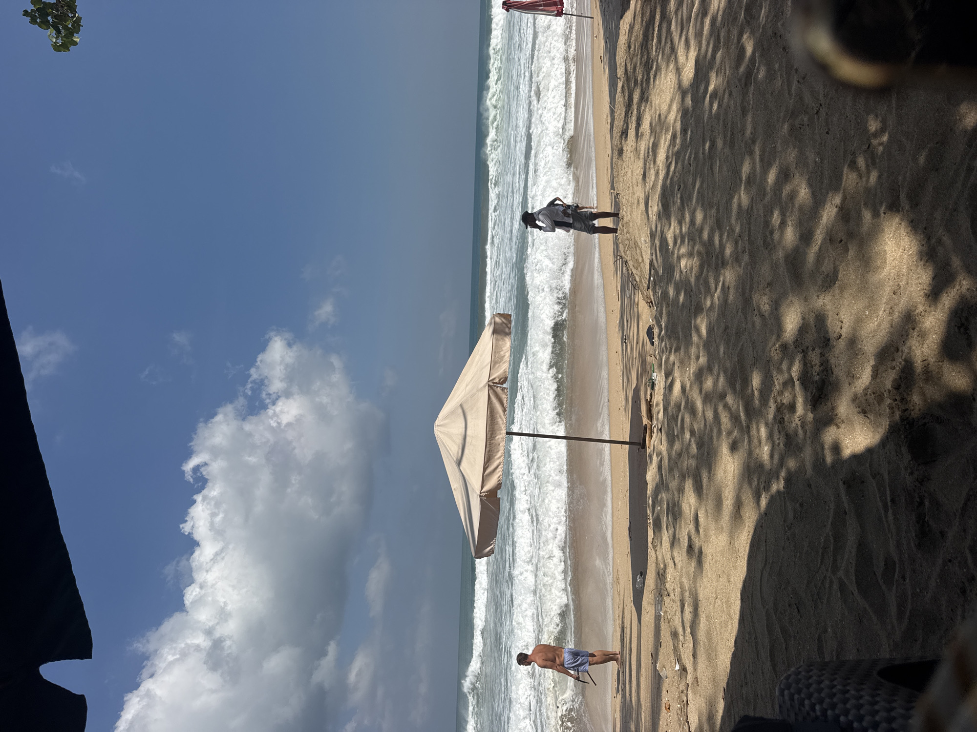 In Kuta, Indonesia, a sunny day unfolds on the beach, with powerful waves crashing onto the shore. The sand is populated by two individuals: a shirtless man in blue shorts stands near the water's edge, holding a long object, while another person, dressed in a white shirt and dark shorts, stands closer to the foreground, engaged in conversation. An umbrella provides shade over the sandy expanse, and trees cast dappled shadows across the scene, indicating a tropical setting. The sky is a vibrant blue, dotted with fluffy white clouds.