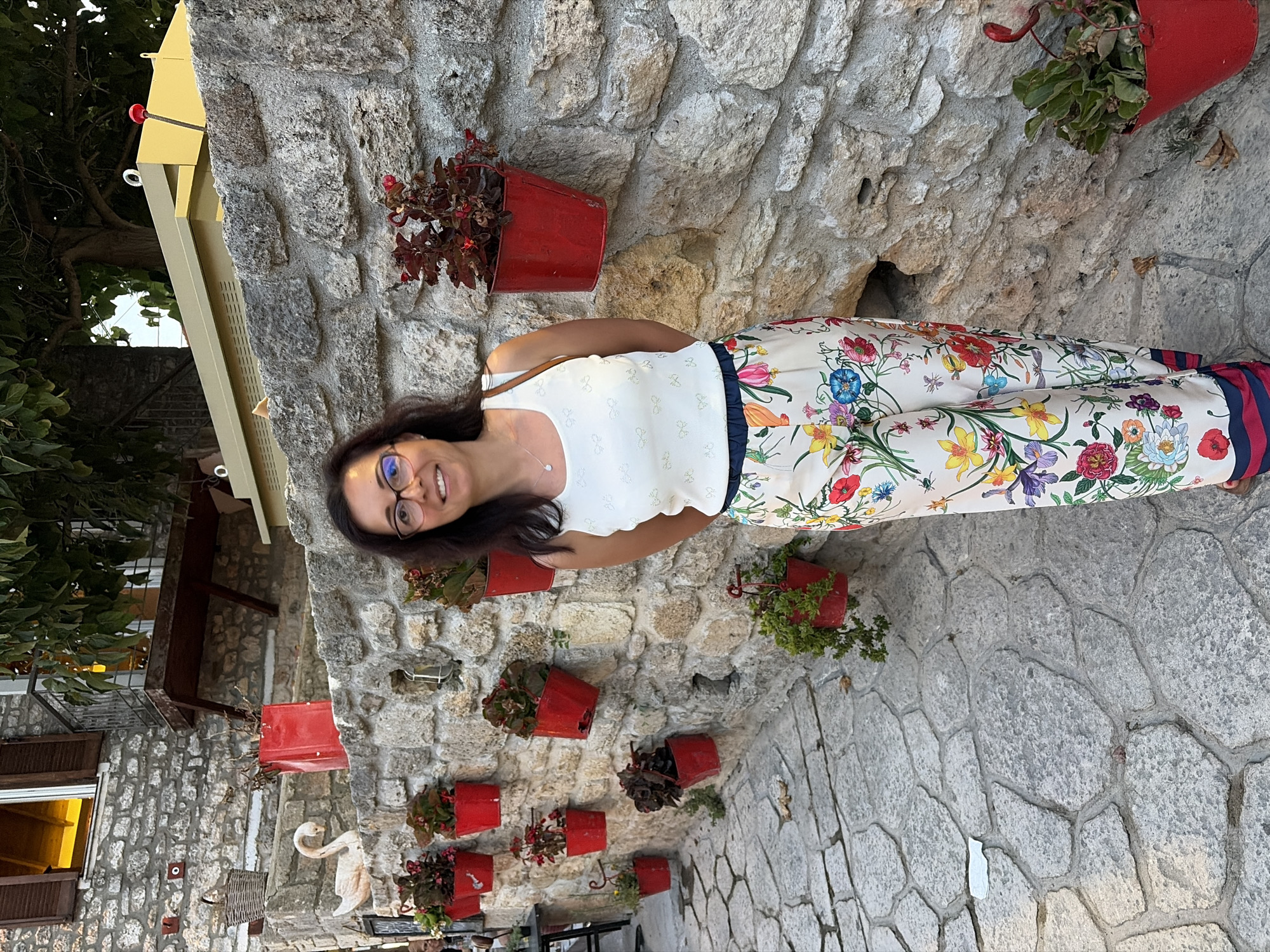 A woman stands against a stone wall adorned with red flower pots. She is wearing a white sleeveless top with a subtle pattern and white pants covered in a vibrant floral print. The woman has dark hair and is wearing glasses. She is smiling and looking towards the camera. The stone wall is rough and textured, with some pots holding green plants and others with darker foliage. The ground is paved with interlocking stones. In the background, a portion of a yellow structure and trees are visible, along with some architectural elements made of stone. The scene suggests an outdoor setting, possibly in a Mediterranean location given the context of Kassandra, Greece. The lighting indicates it is daytime.