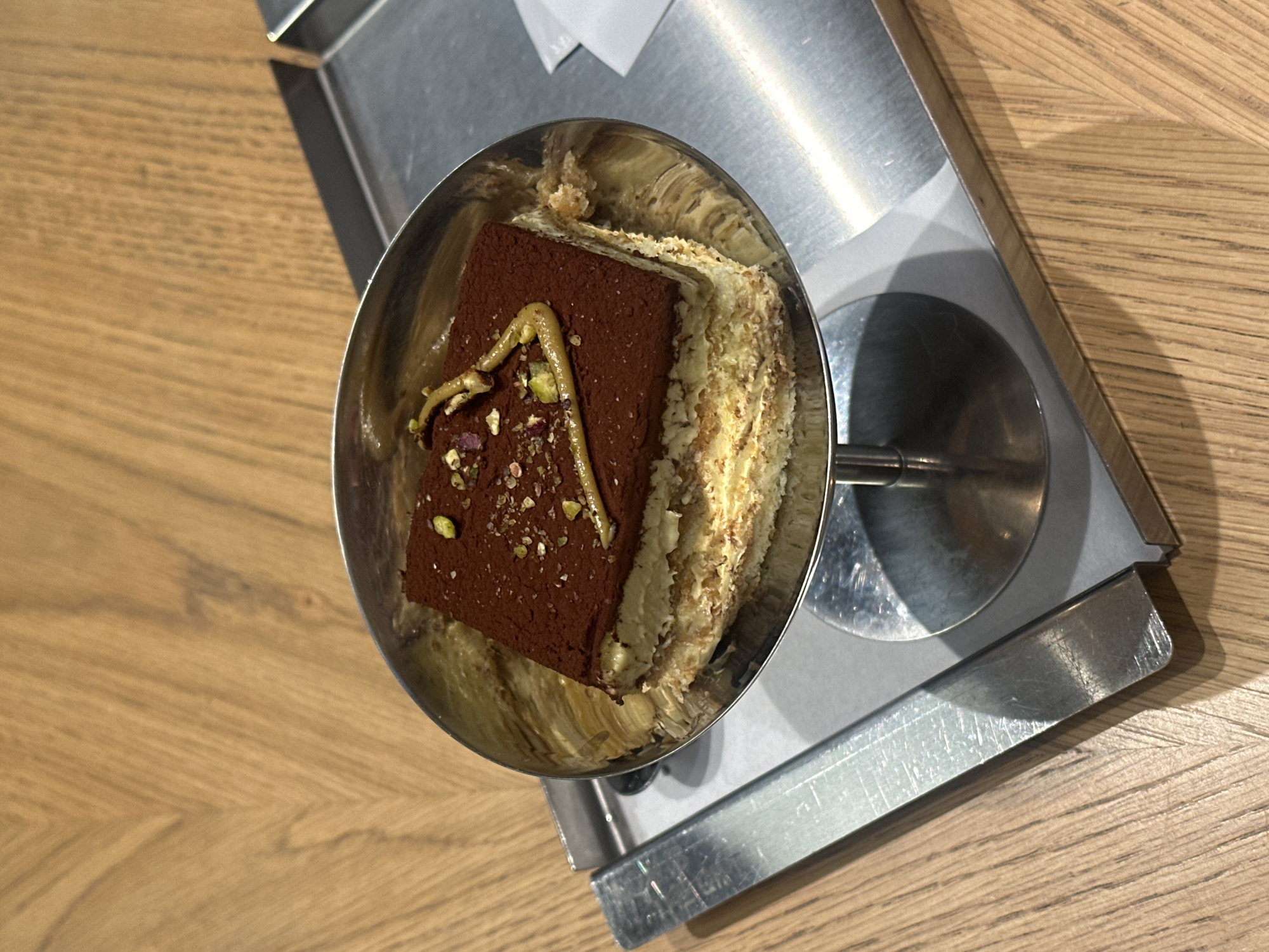 The image captures a dessert served in a metal cup, positioned on a metallic tray that sits on a wooden table. The dessert features a chocolate-brown square topping dusted with what appears to be pistachio and possibly rose petal sprinkles, drizzled with a light yellow sauce. Beneath the topping, there are layers of what looks like cream and pastry. The lighting suggests an indoor setting. This image was taken in Yerevan, Armenia.