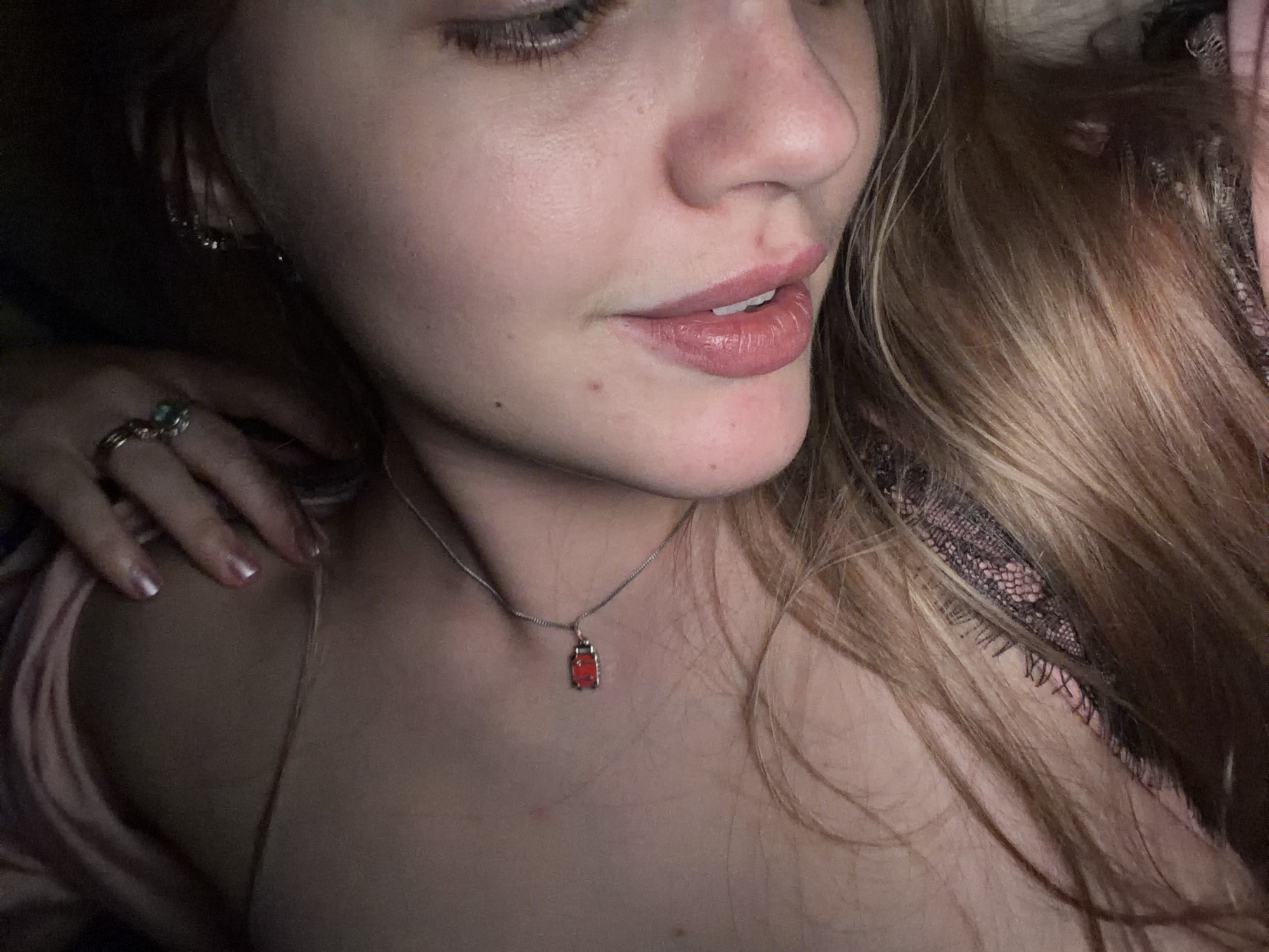 The image shows a close-up of a woman's face and upper body. She has light skin, blonde hair, and is wearing a silver necklace with a small red pendant. Her lips are slightly parted. A hand with rings on the fingers rests on her shoulder. Part of her clothing appears to be lace. There is no information available to indicate this image was taken in Большая Шильна, Russia.